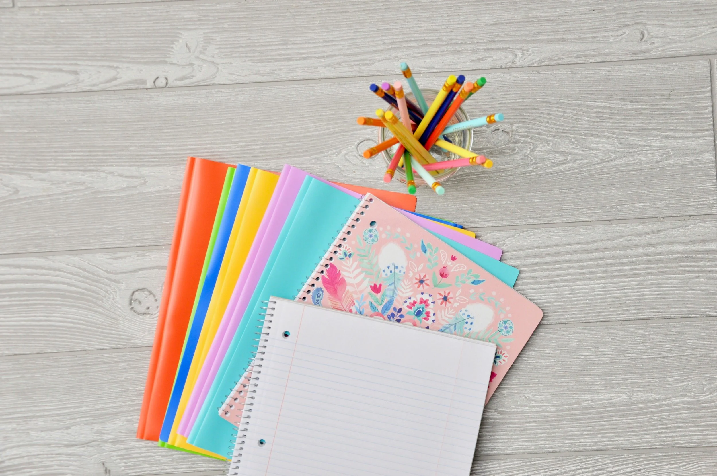 6 school folders in a variety of colors, two notebooks, and a cup of colorful pencils all sitting on a desk.
