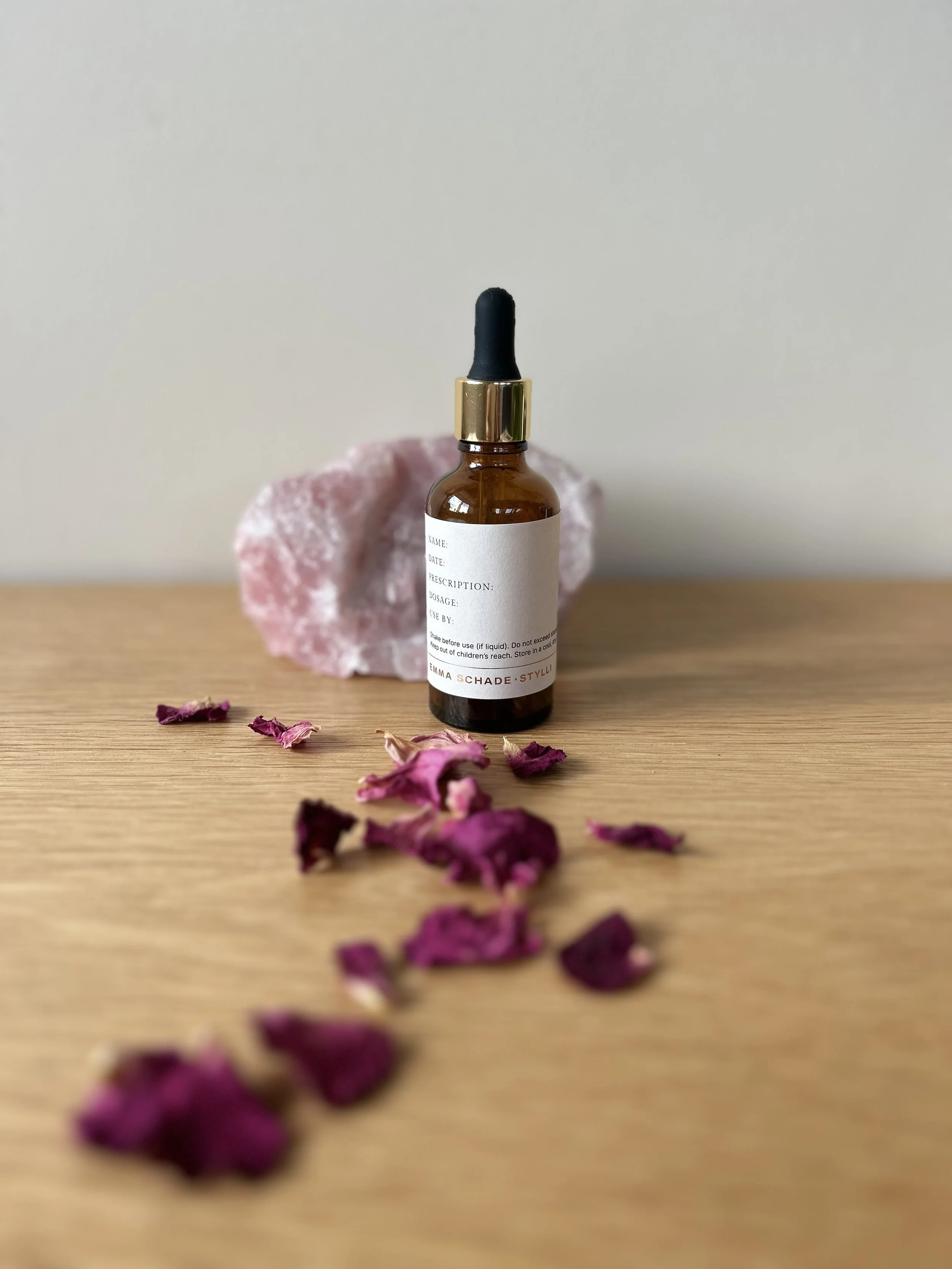 An amber dropper bottle with a custom prescription label by Emma Schade-Stylli, resting beside a raw rose quartz crystal and a pathway of dried pink rose petals on a light wood table.