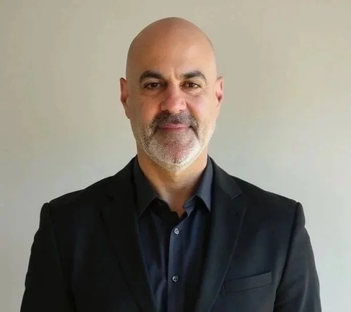 A middle-aged man with a bald head and gray beard wearing a black suit and black shirt, standing against a plain light-colored wall.