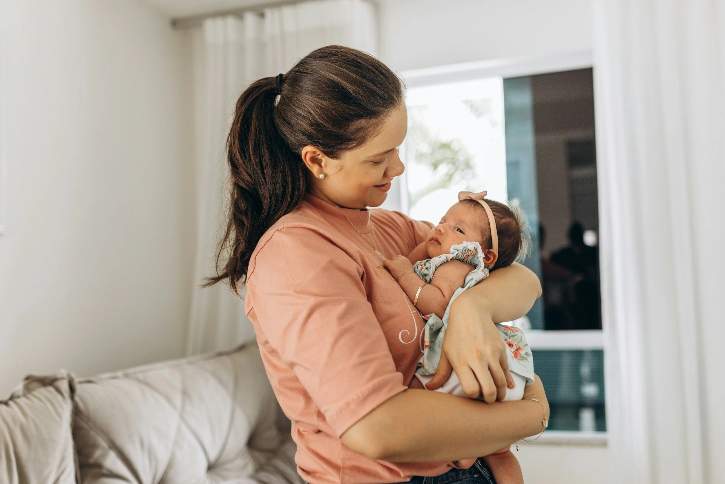 Mother in a pink top gently holding her newborn baby girl in a bright living room, illustrating the new parent support group for postpartum mothers