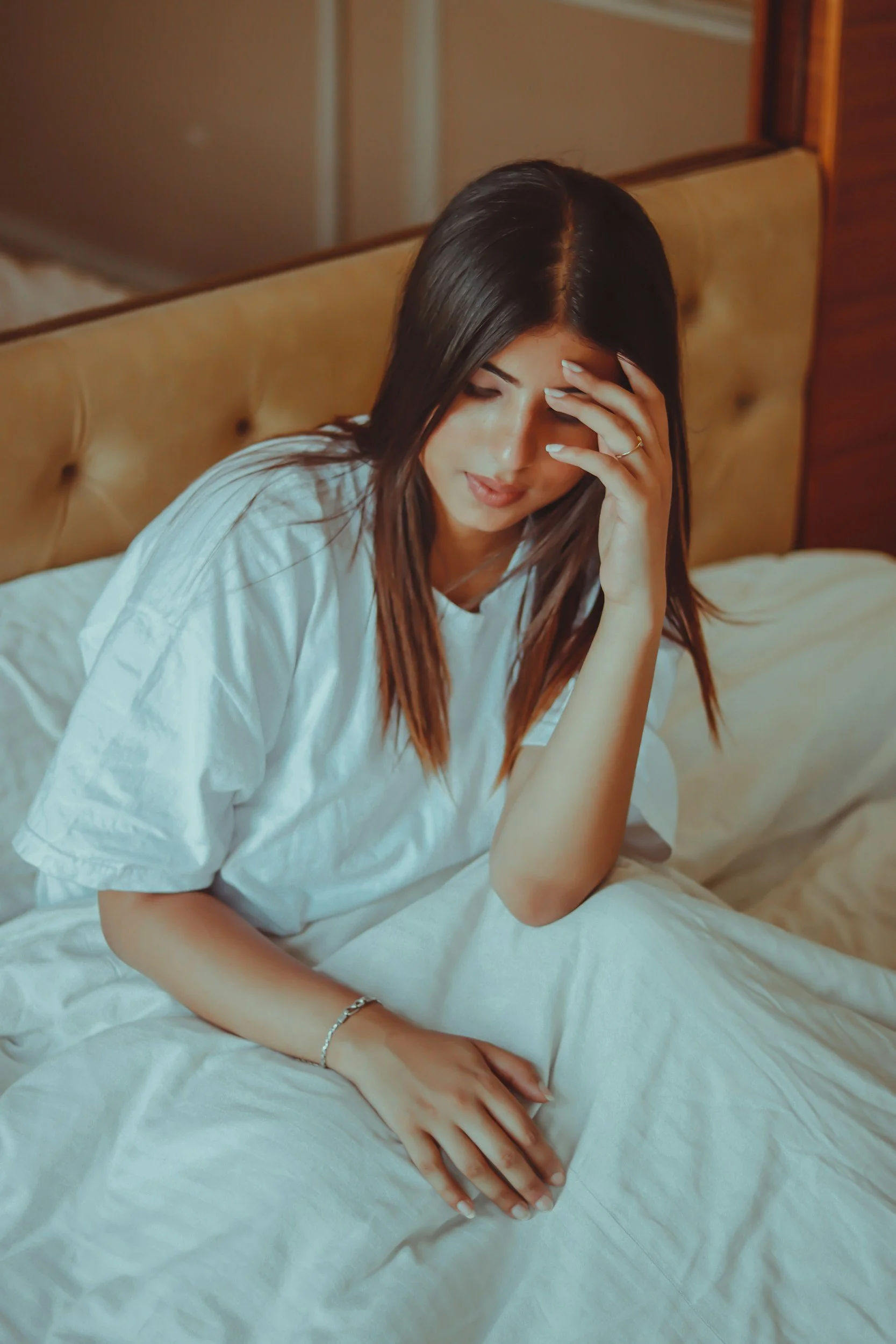Woman in white sitting on a bed with her hand on her forehead in quiet sorrow, illustrating the early pregnancy loss support group