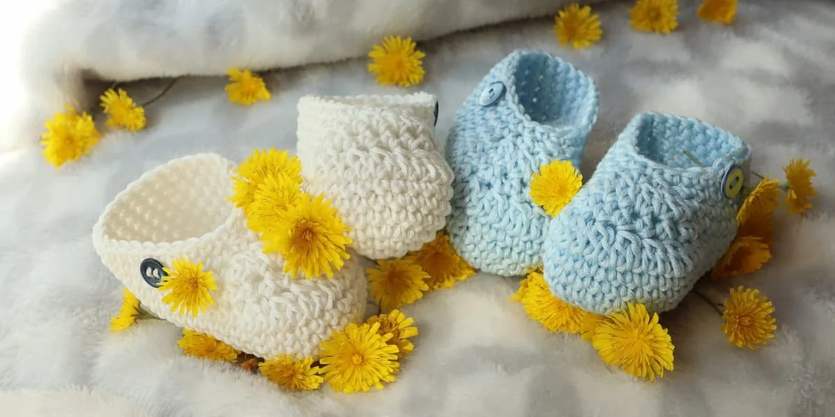 White and blue knitted baby booties surrounded by yellow flowers on a soft white blanket, symbolizing remembrance and love for babies lost during pregnancy or infancy.