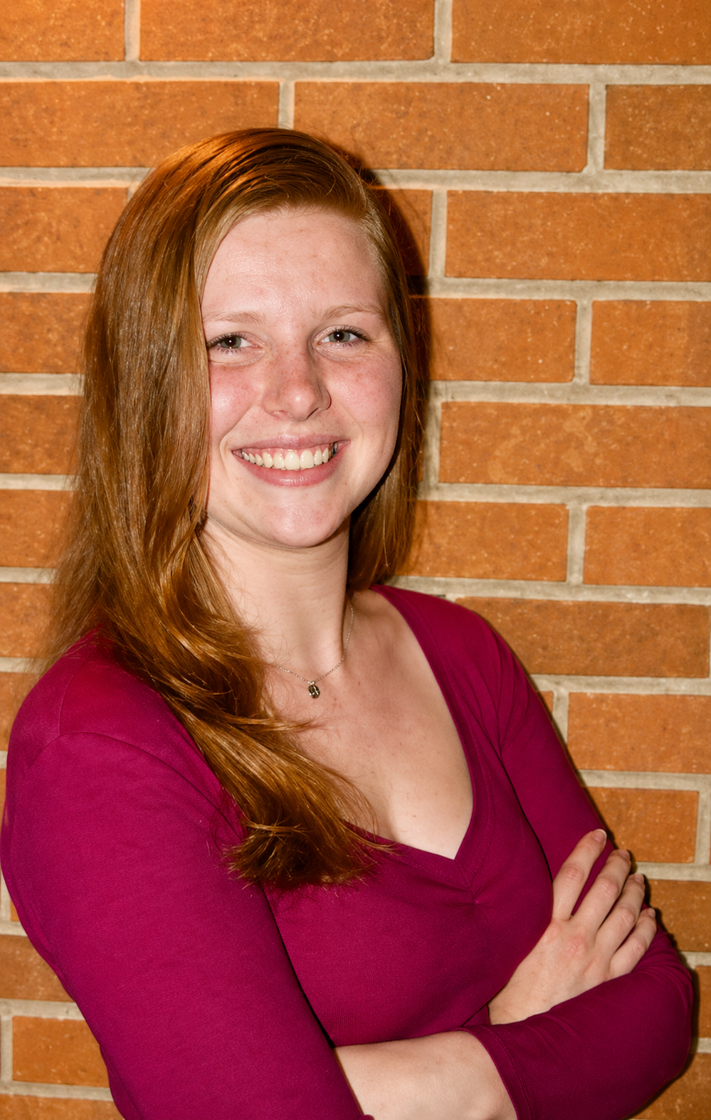 Portrait of Haleigh Champagne, Outreach Coordinator at Hearth Counseling & Consulting, smiling with arms crossed, wearing a magenta top, standing in front of a brick wall.
