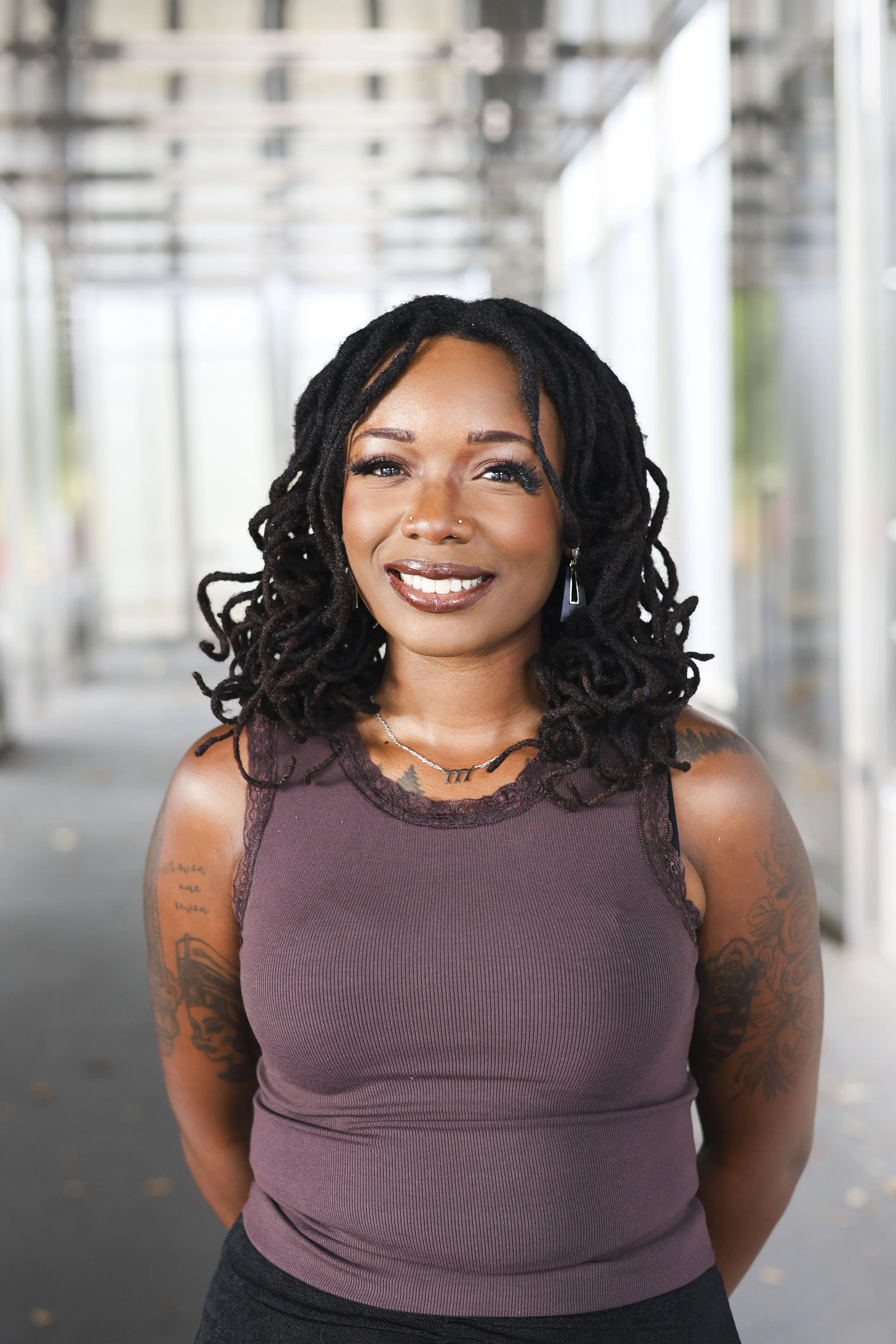 Smiling person with shoulder-length black locs wearing a sleeveless brown top in a bright, modern hallway.