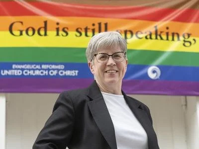 A smiling woman with short gray hair and glasses standing in front of a rainbow pride flag with the words 'God is still speaking' and 'BIBLICAL, REFORMED UNITED CHURCH OF CHRIST' written on it.