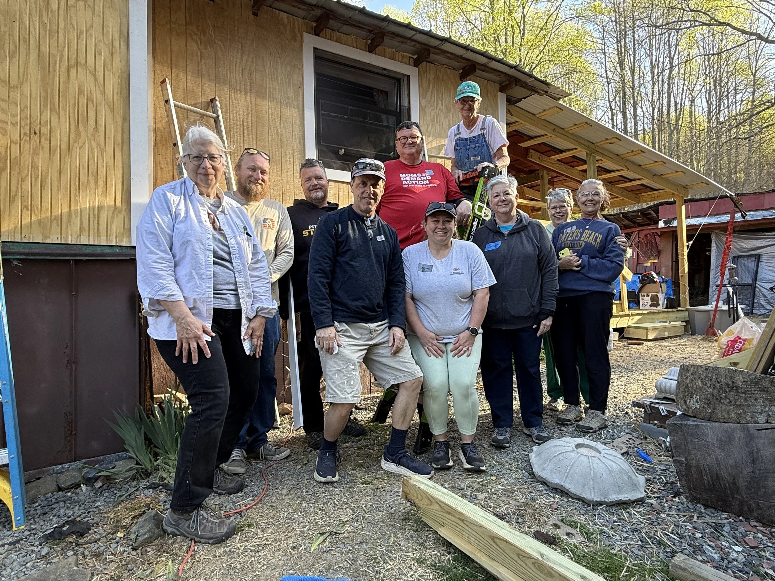 Group of people standing outside a house under construction, some wearing casual clothes and safety gear, smiling at the camera.