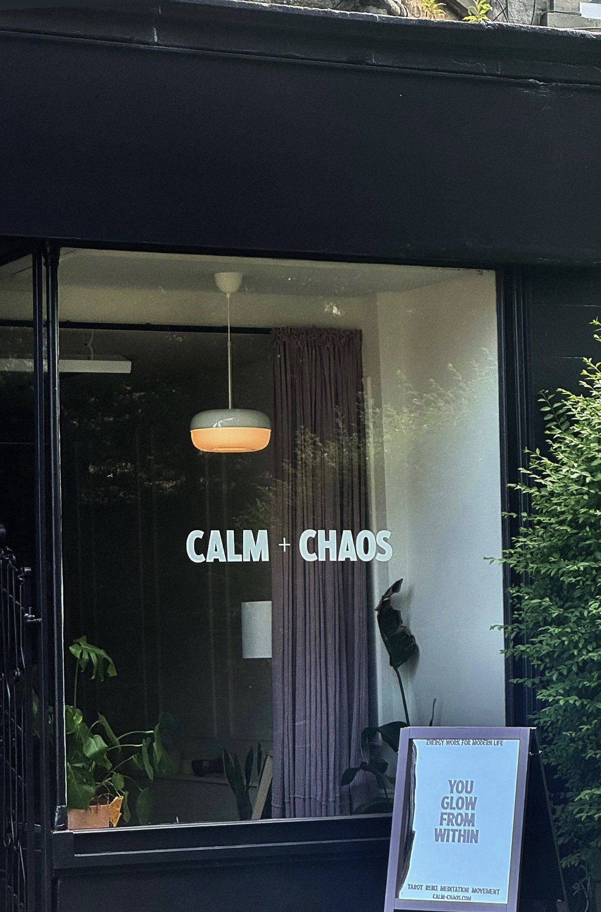 A window with the text 'CALM + CHAOS' and a hanging light inside the room. There's a purple curtain, some plants, and a standing sign outside with the text 'YOU GLOW FROM WITHIN'.