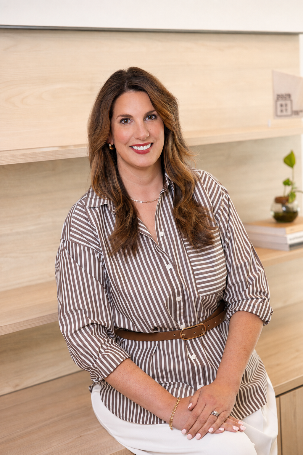 A woman with long brown hair sitting on a wooden surface, smiling at the camera, wearing a brown and white striped blouse and white pants, with a small plant and books in the background.