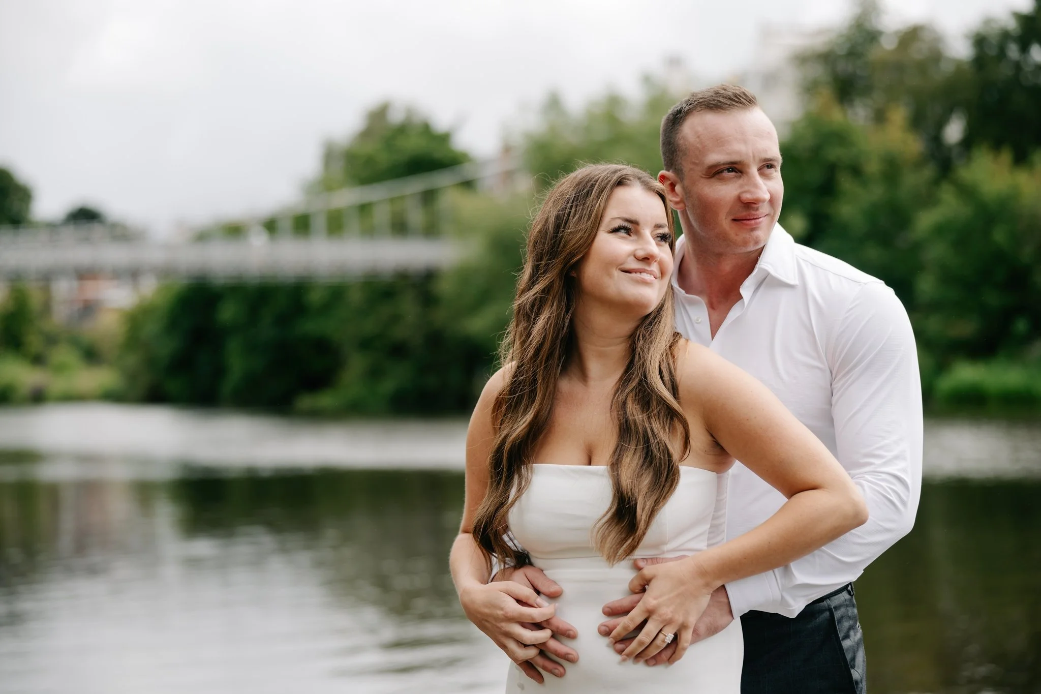 A couple stands close together outdoors near water, with the man embracing the woman from behind. The woman, wearing a white dress, has long brown hair and is smiling softly, looking into the distance. The man is dressed in a white shirt and dark pants, looking off to the side with a gentle expression. Green trees and a bridge are visible in the background.