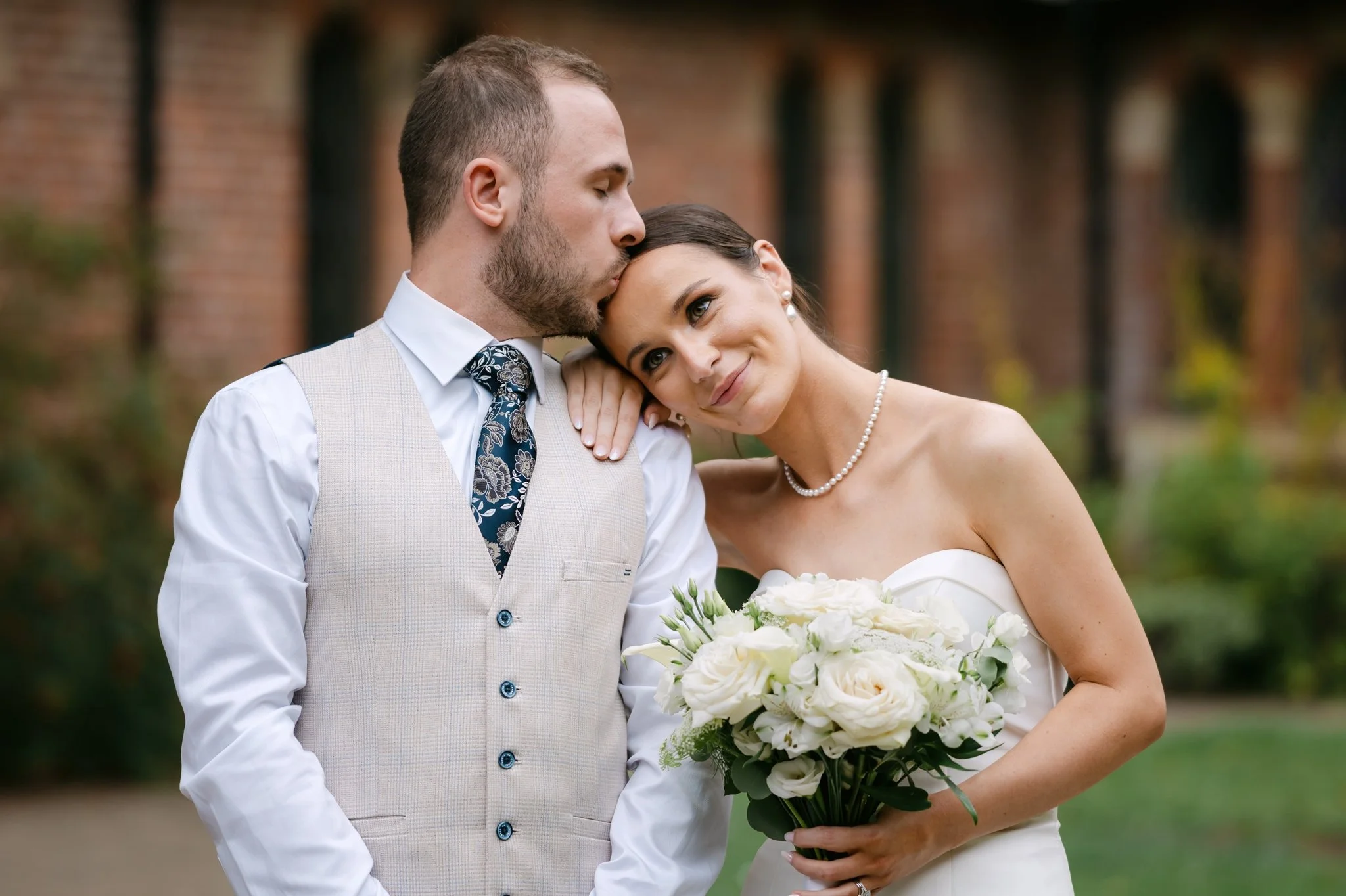 A newlywed couple outdoors, the groom gently kisses the bride's forehead while she holds a bouquet of white flowers, smiling softly, with a blurred background of trees and a brick building.