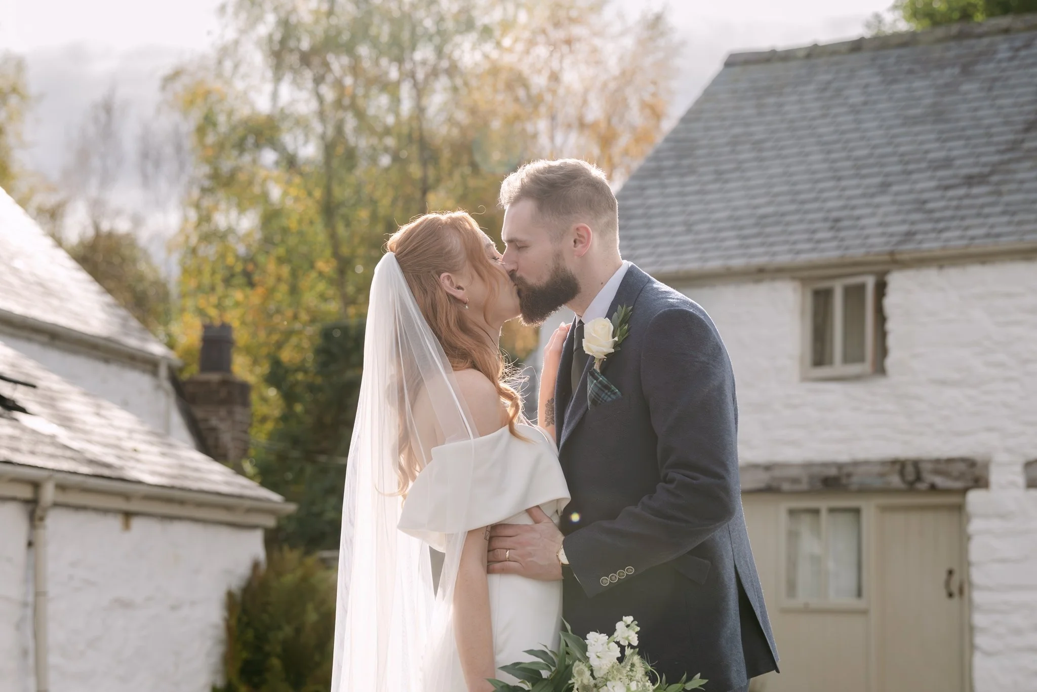 A bride and groom share a kiss outdoors during their wedding ceremony, with a rustic white building and trees with autumn foliage in the background.