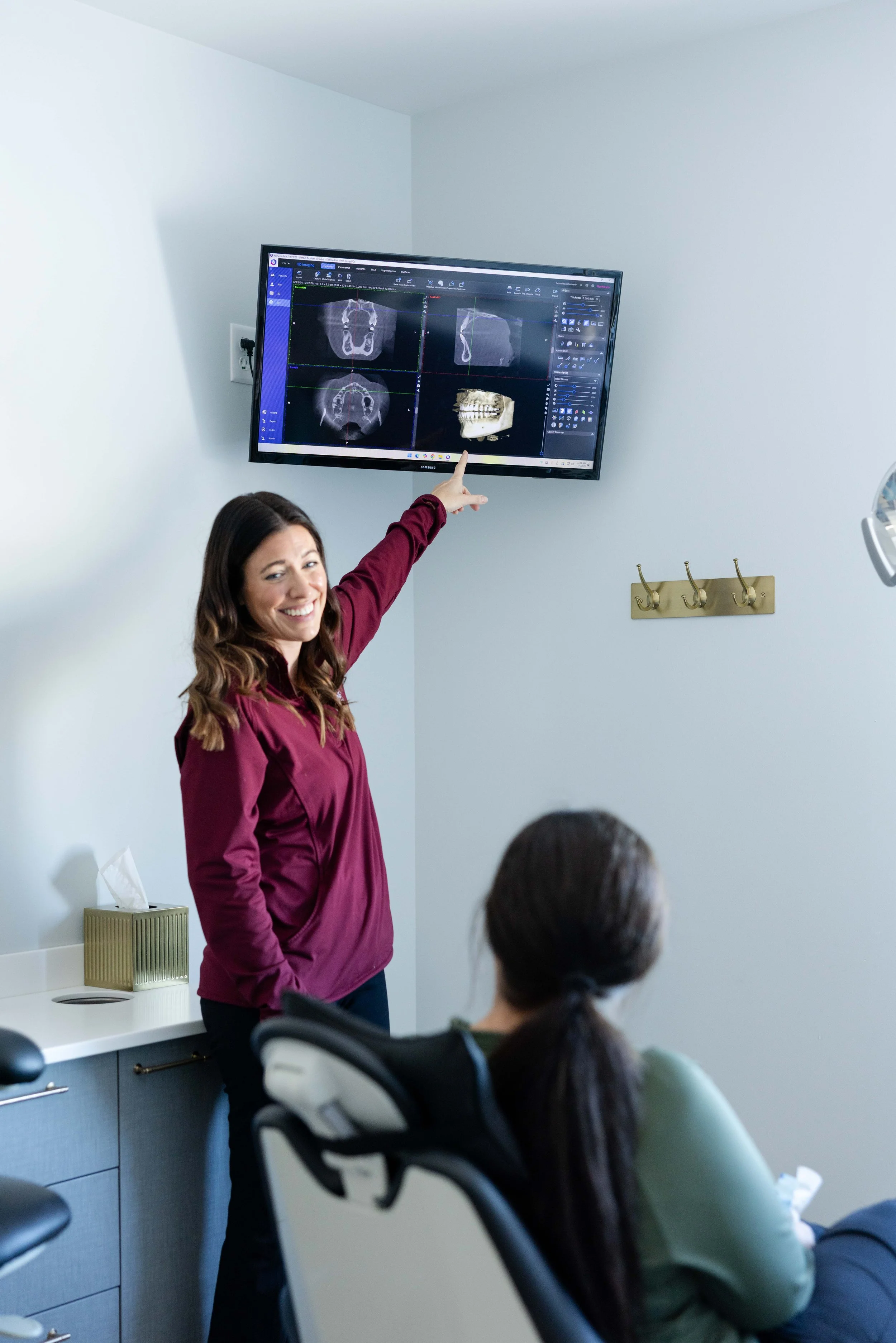 A woman in a maroon blouse is pointing to dental X-ray images displayed on a wall-mounted monitor in a dental office, while a patient seated in a dental chair listens.