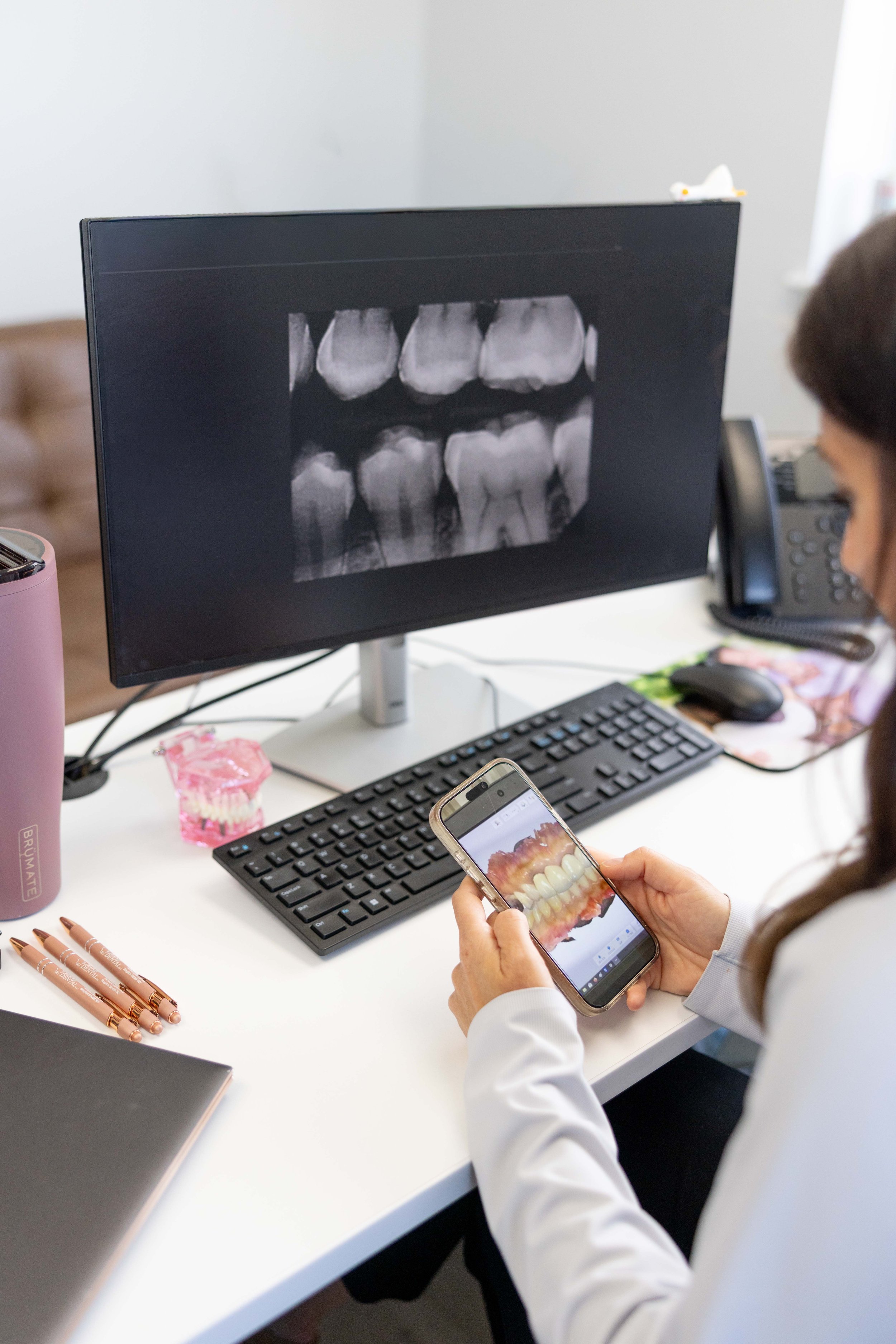 Person in a white lab coat viewing a 3D dental model on a smartphone, with a computer monitor displaying a dental X-ray in the background.