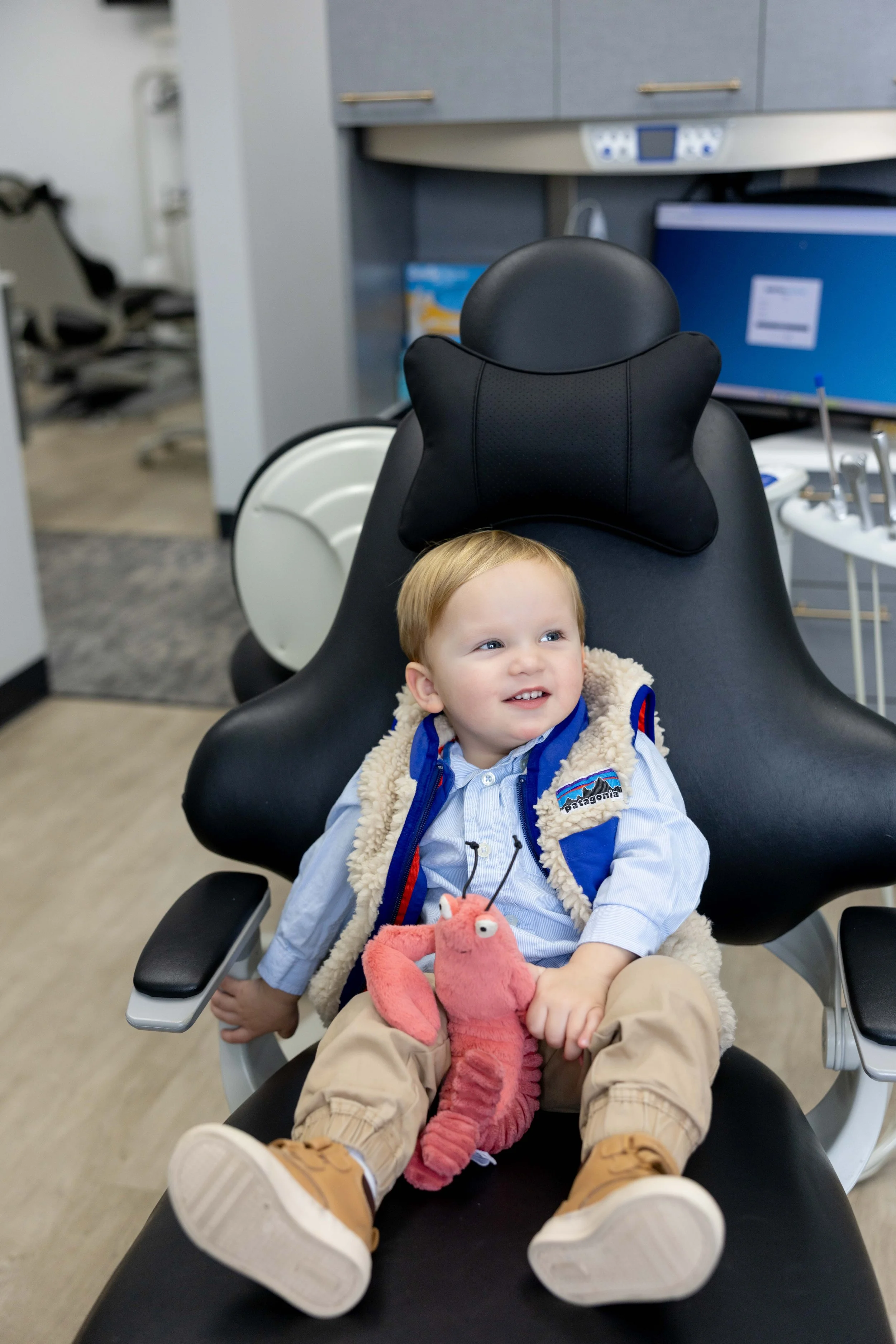 A young boy with blonde hair sitting in a dentist's chair with a black headrest and a black cushion shaped like a butterfly behind his head. He is holding a pink plush toy of the cartoon character from the Pinkfong video. The boy is smiling and looking to his left. The background shows dental equipment, a computer monitor, and cabinets in a dental office.