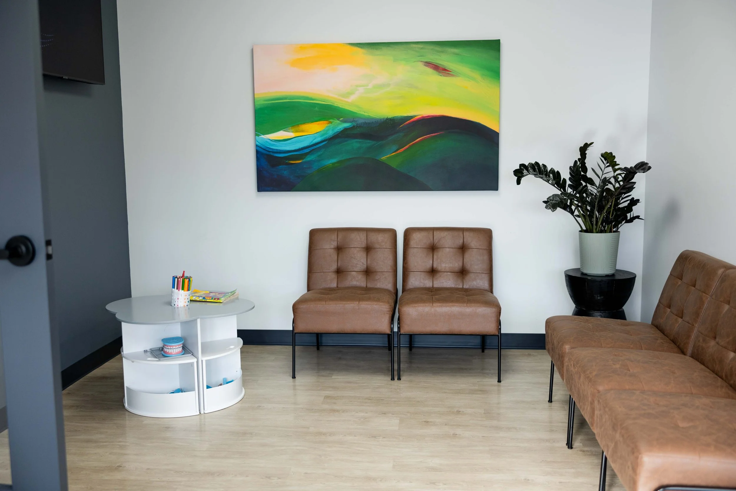 Waiting room with brown leather chairs, a white coffee table with coloring supplies, a large abstract painting with green, yellow, and blue colors on a white wall, and a tall plant in a pot.
