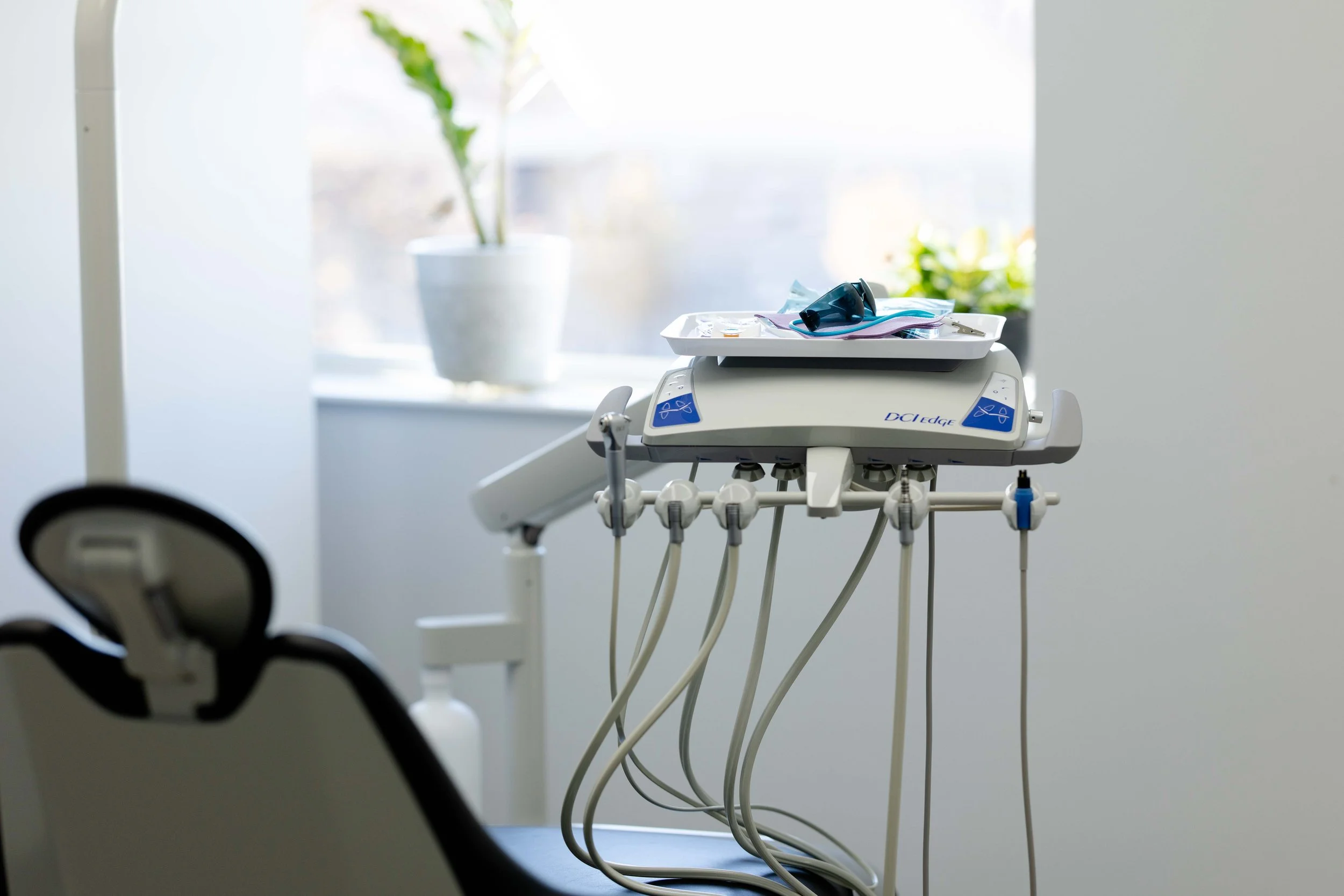 A dental treatment room with dental equipment including a tray with protective glasses, a face mask, and dental tools on a mobile cart near a window with potted plants.