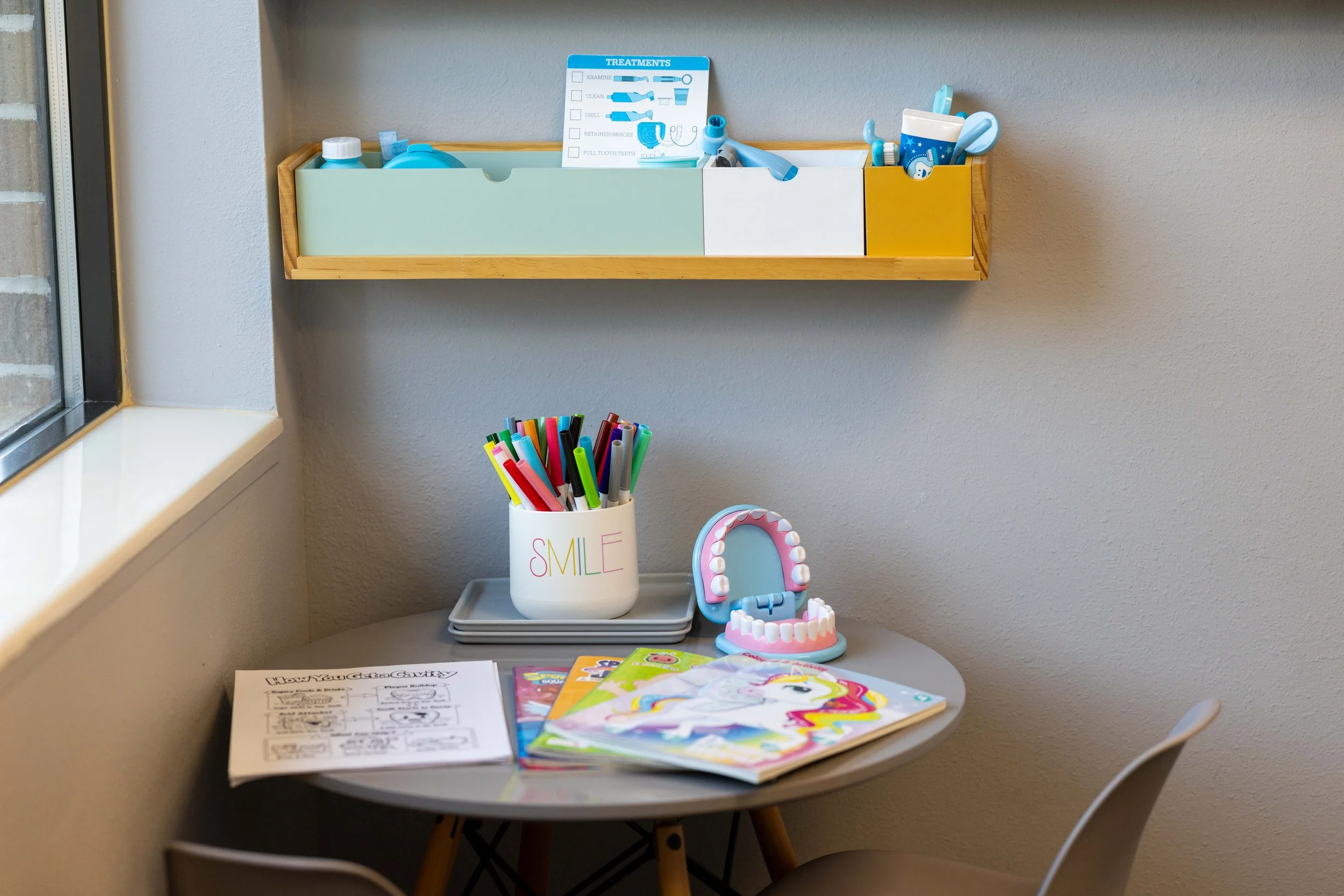 Small round table with coloring books, a container of colored markers, and a dental mirror. On the wall above, a wooden shelf holding a diagram of dental treatments, saliva bottles, and dental tools. Outside the window, part of a brick wall is visible.