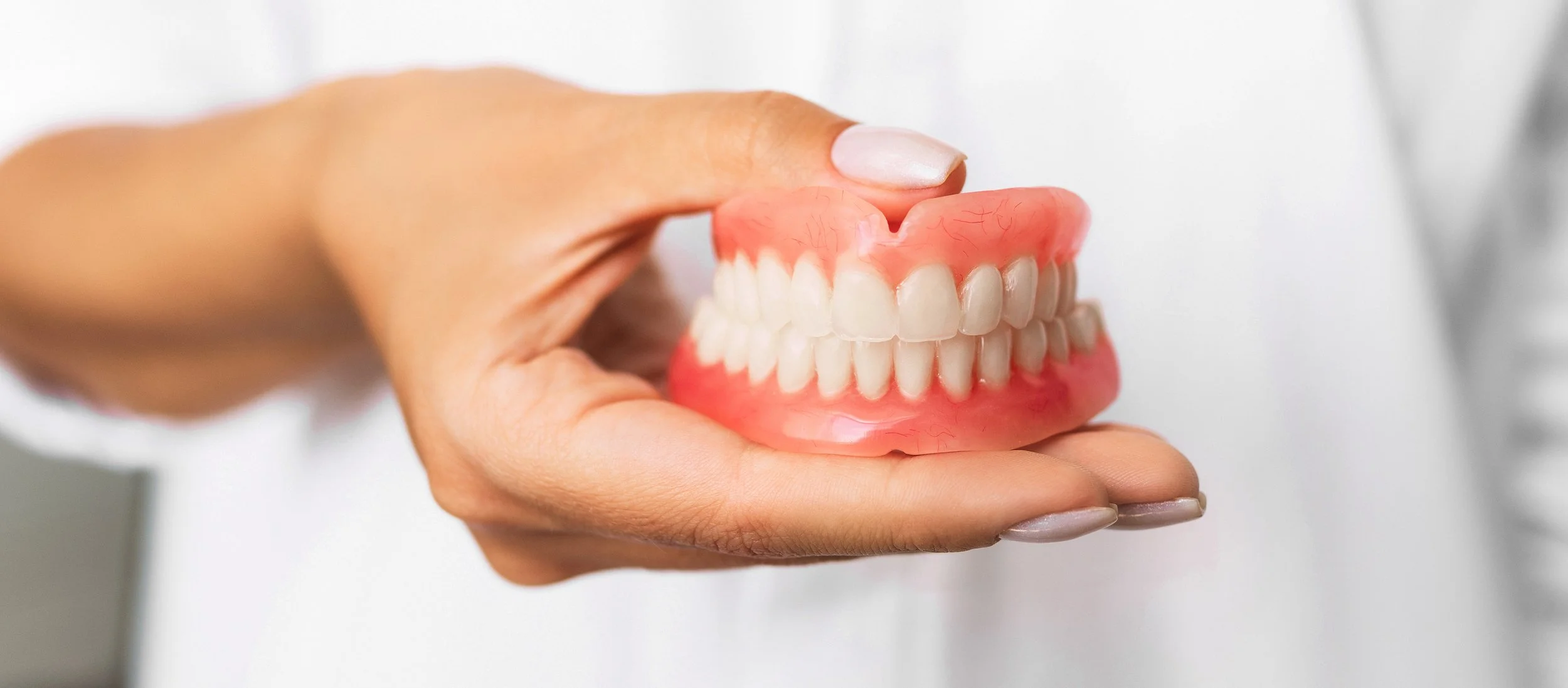 A hand holding a set of dental dentures with white teeth and pink gums.
