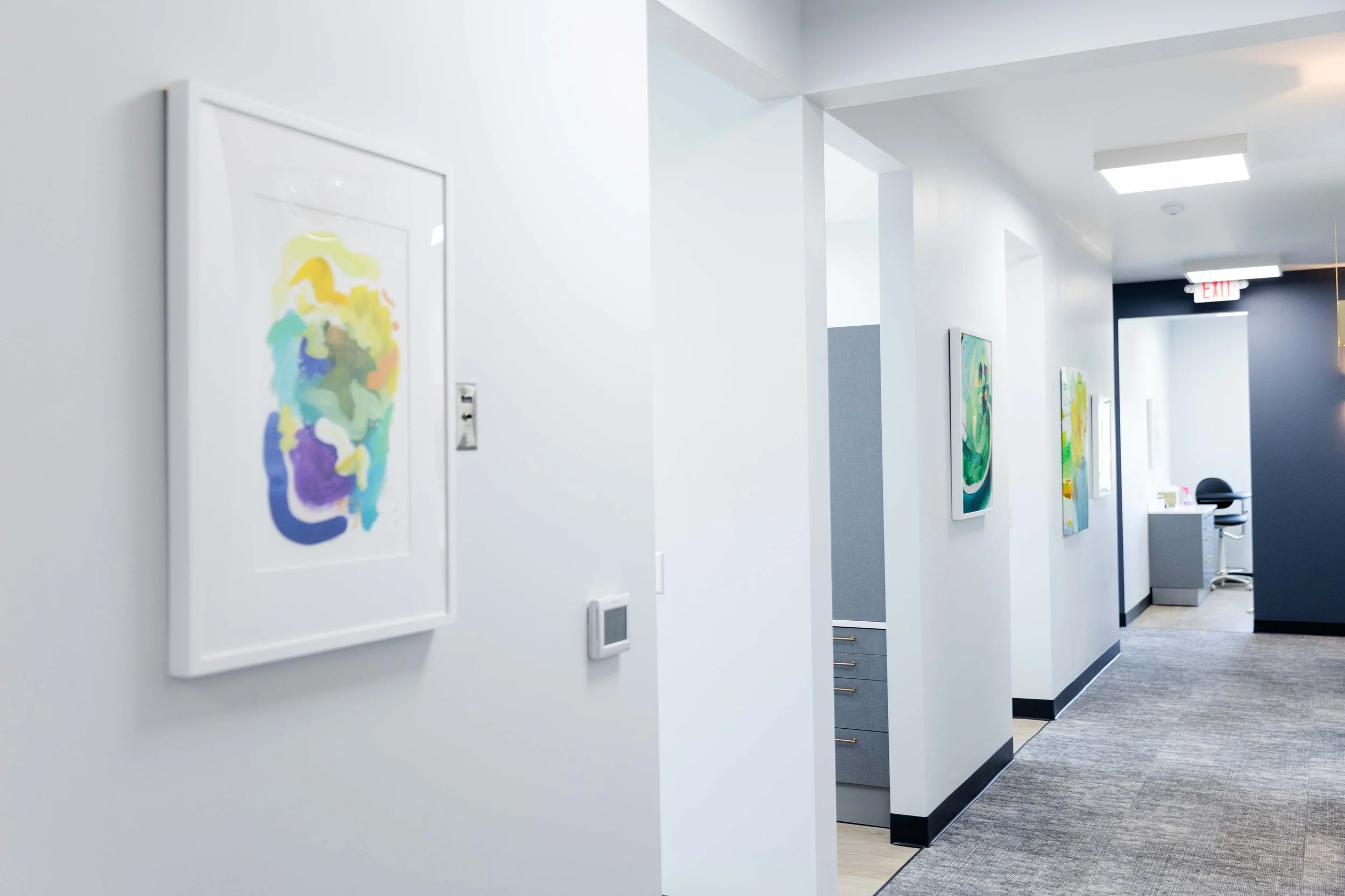 White corridor with colorful abstract paintings hanging on the walls, leading to a reception area with a dark blue wall, an exit sign, and a desk with a black chair.