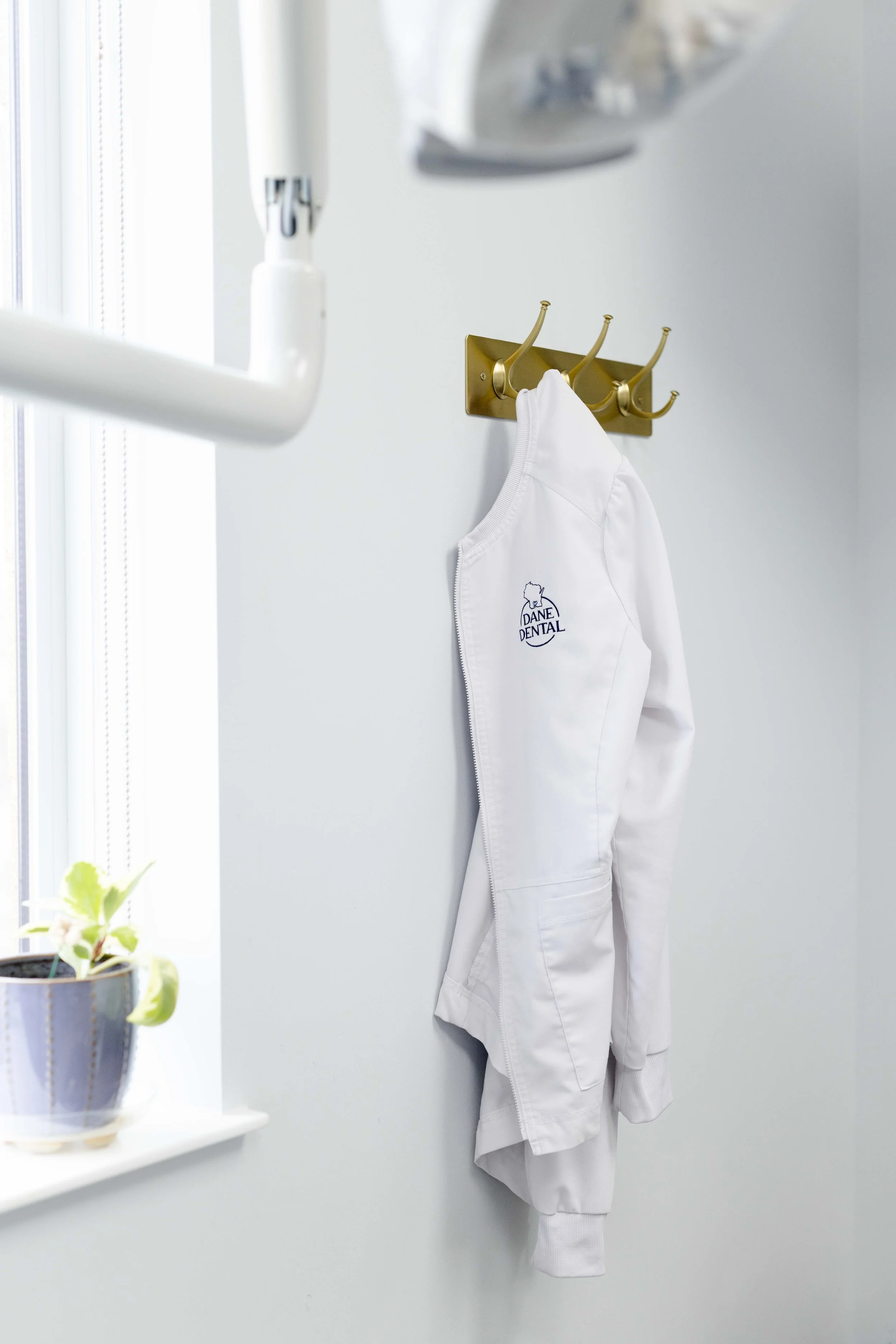 White dental lab coat hanging on a gold wall hook in front of a white wall, with a window and potted plant in the background.