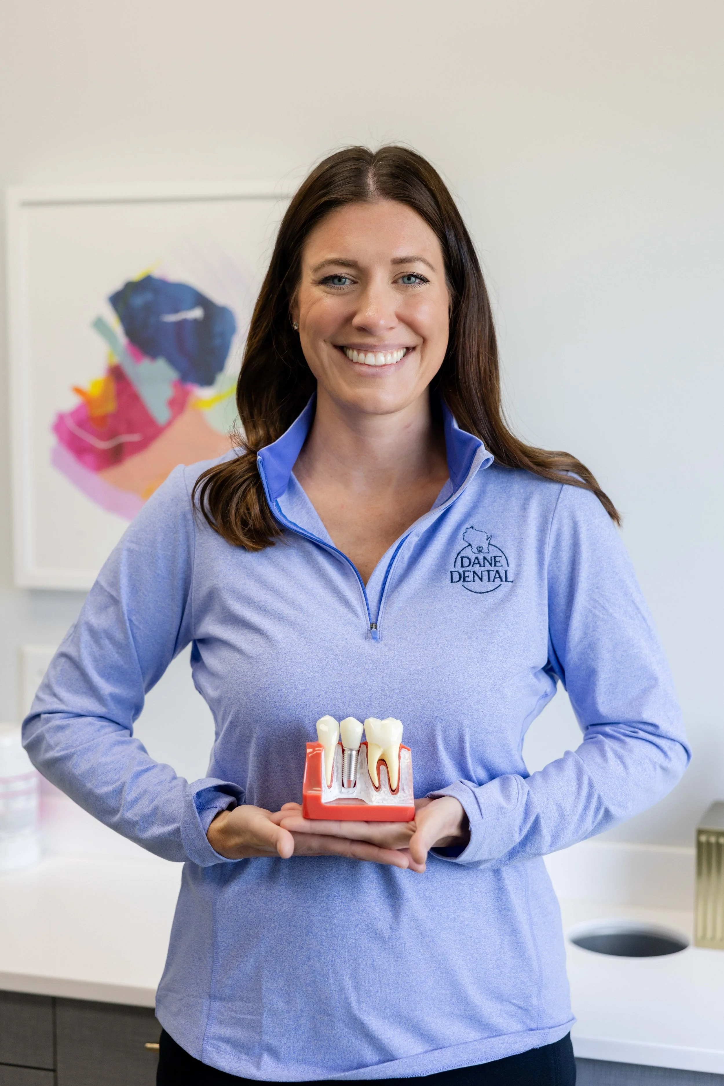 A woman in a blue dental uniform smiling and holding a dental model with teeth, standing in a dental office with colorful artwork on the wall.
