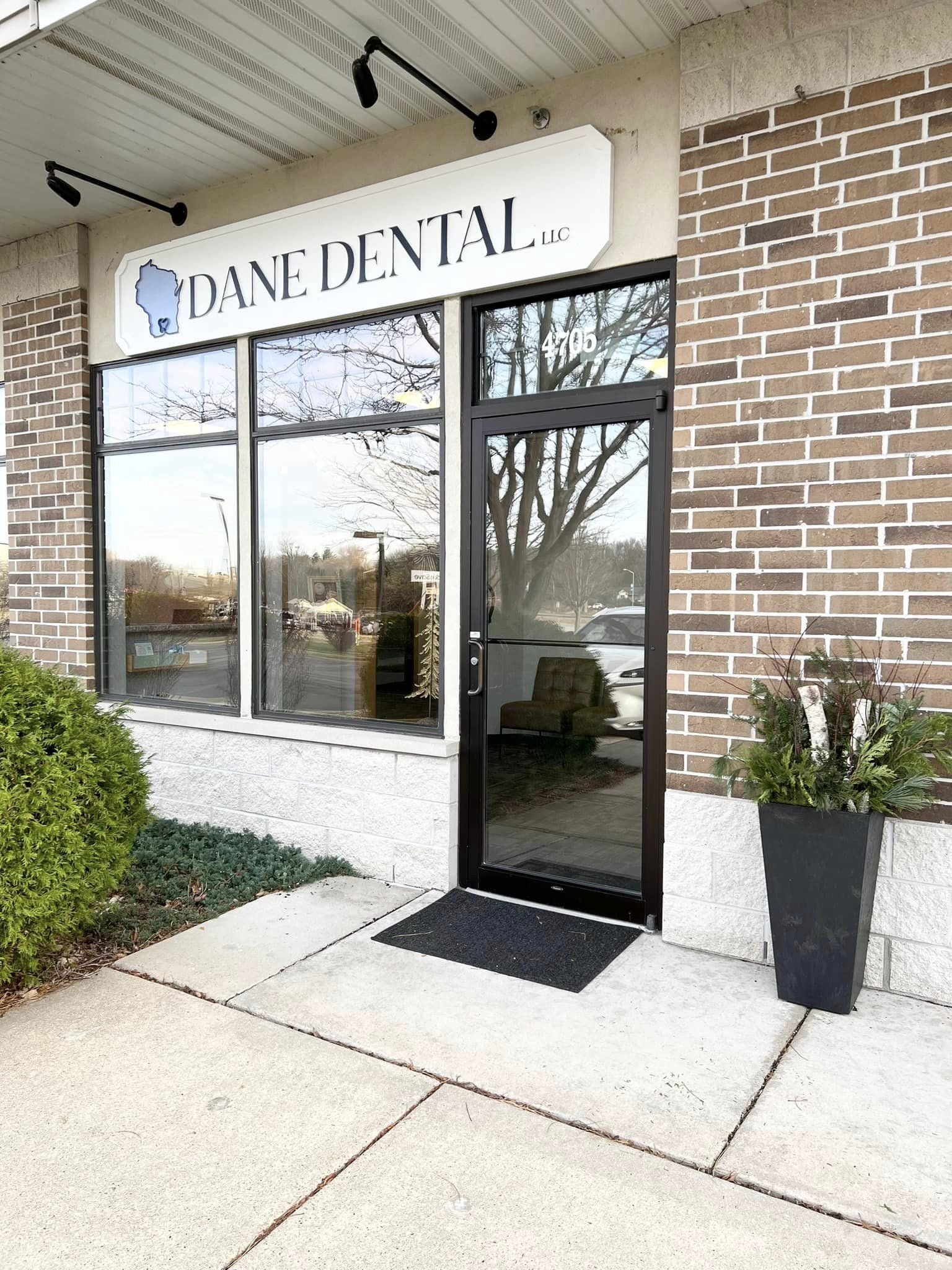 Exterior view of a dental office with a sign that reads 'Dane Dental LLC' and includes a map outline of Wisconsin. The entrance has glass doors with a black frame, a potted plant to the right, and a small bush and concrete walkway outside.