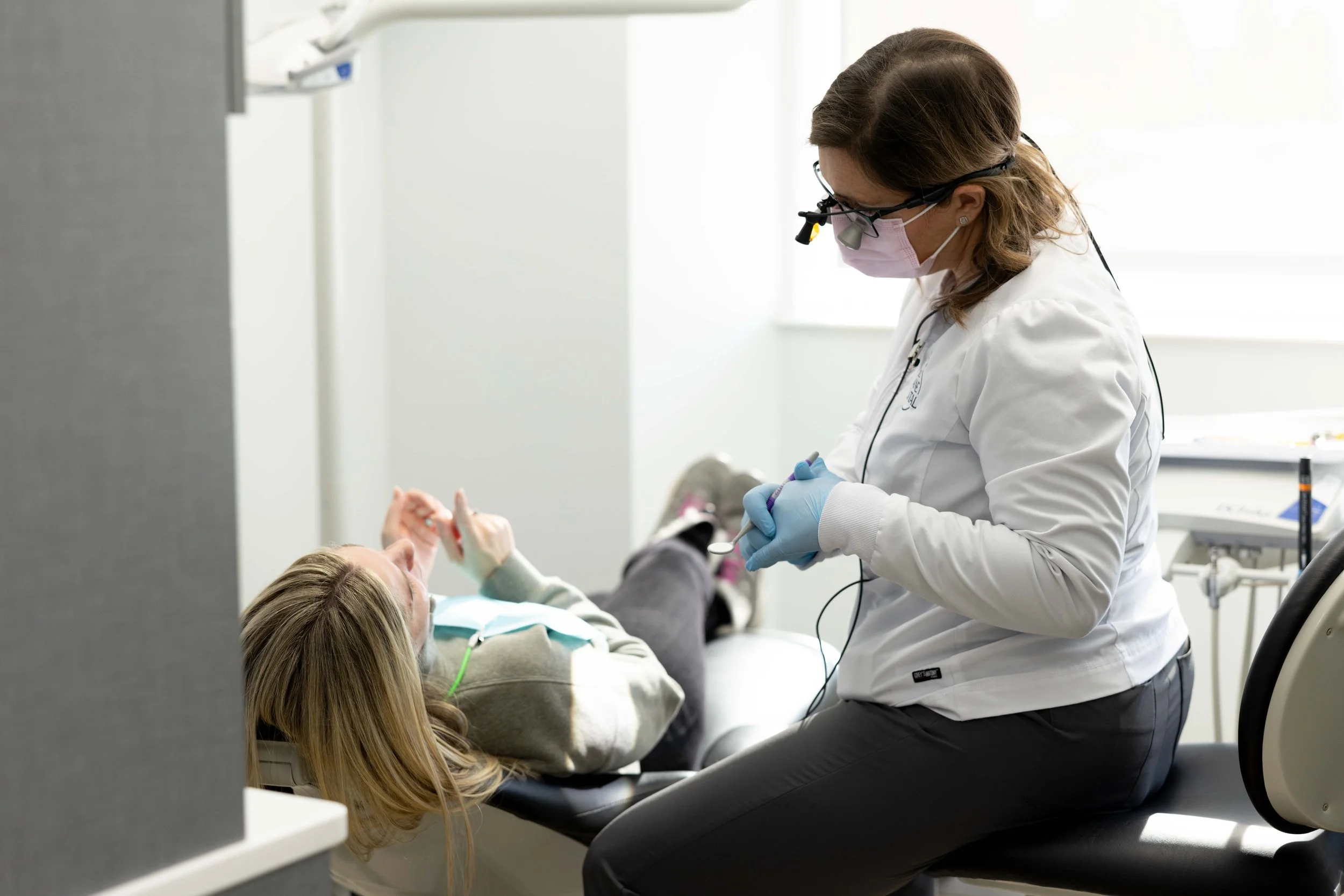 A healthcare professional wearing glasses, a pink face mask, and gloves, attending to a patient lying on a dental chair in a bright clinic. The patient has long blonde hair.