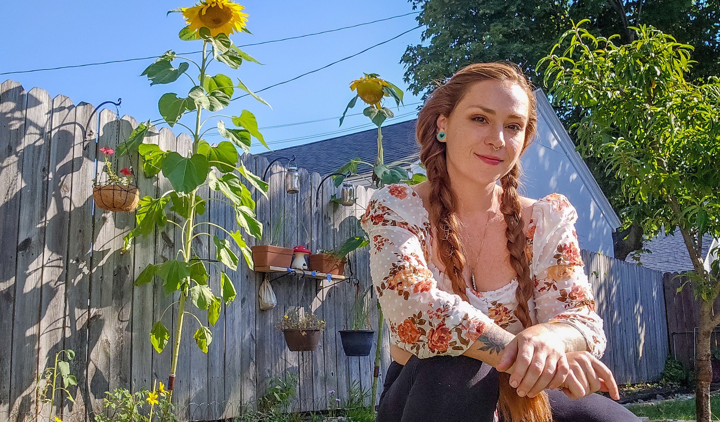A young woman with long red hair in braids, wearing a floral dress, sitting outdoors next to a wooden fence decorated with plants and hanging jars, with green trees and clear blue sky in the background.