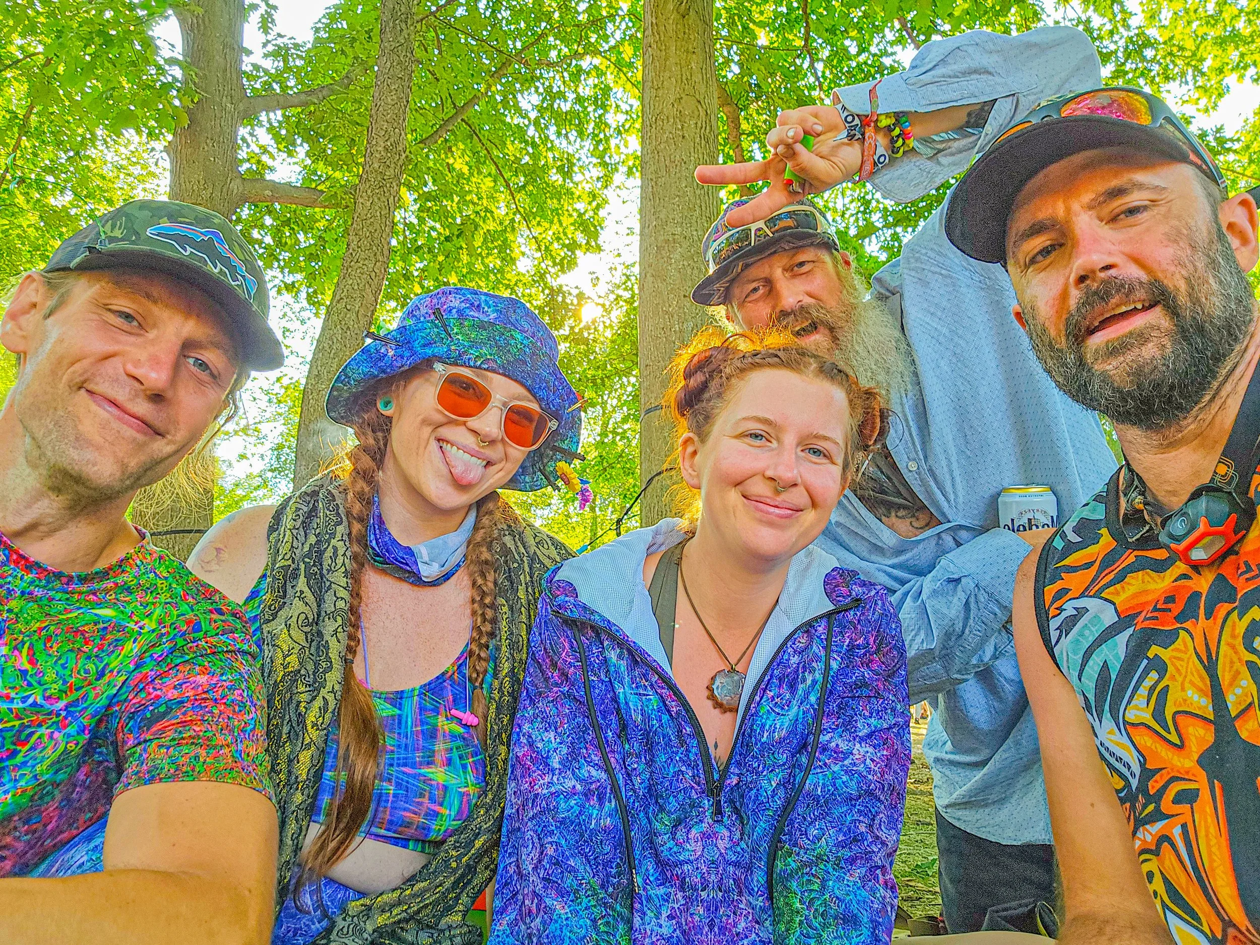 A group of people outdoors at Electric Forest, wearing Morgan Starfox designed clothing, smiling and posing for a selfie. They are surrounded by green trees and sunlight streaming through the leaves.