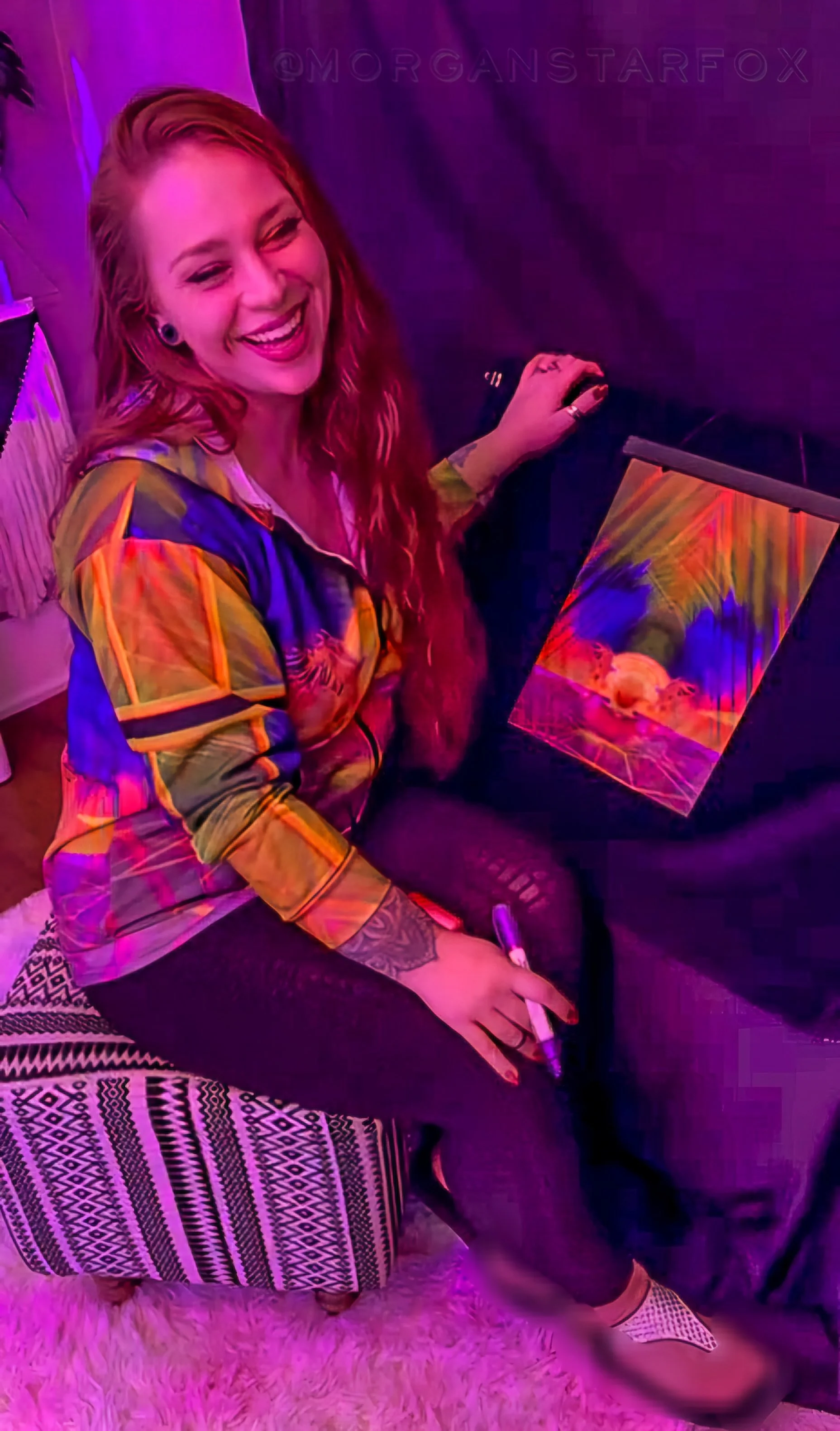 A woman with long, red hair sitting on a patterned ottoman, smiling, wearing a colorful jacket, holding a white marker, with a colorful artwork in front of her, in a room with purple lighting.