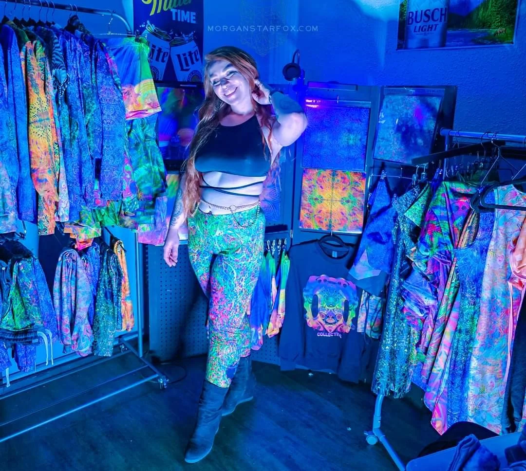 A woman with long red hair smiling, wearing a black top with cutouts, colorful patterned pants, and black boots in a room with neon lighting surrounded by tie-dye and psychedelic clothing.