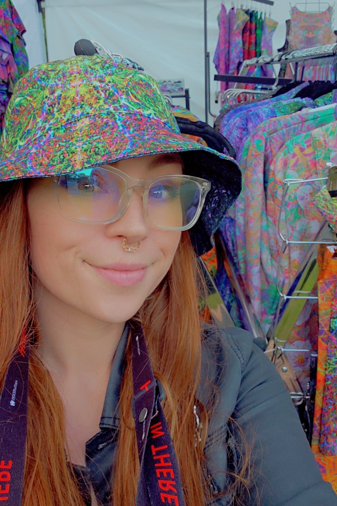 A young woman with long red hair, wearing glasses, a colorful hat, and a septum piercing, taking a selfie in front of a display of tie-dye clothing.
