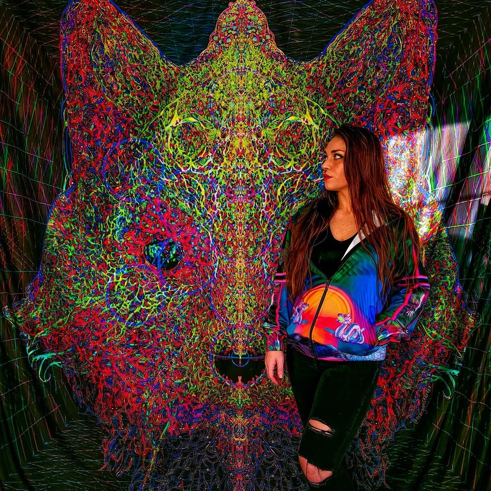 A woman with long red hair in a colorful, patterned jacket stands in front of a vibrant, psychedelic mural of a fox's face made up of intricate multicolored lines and shapes.