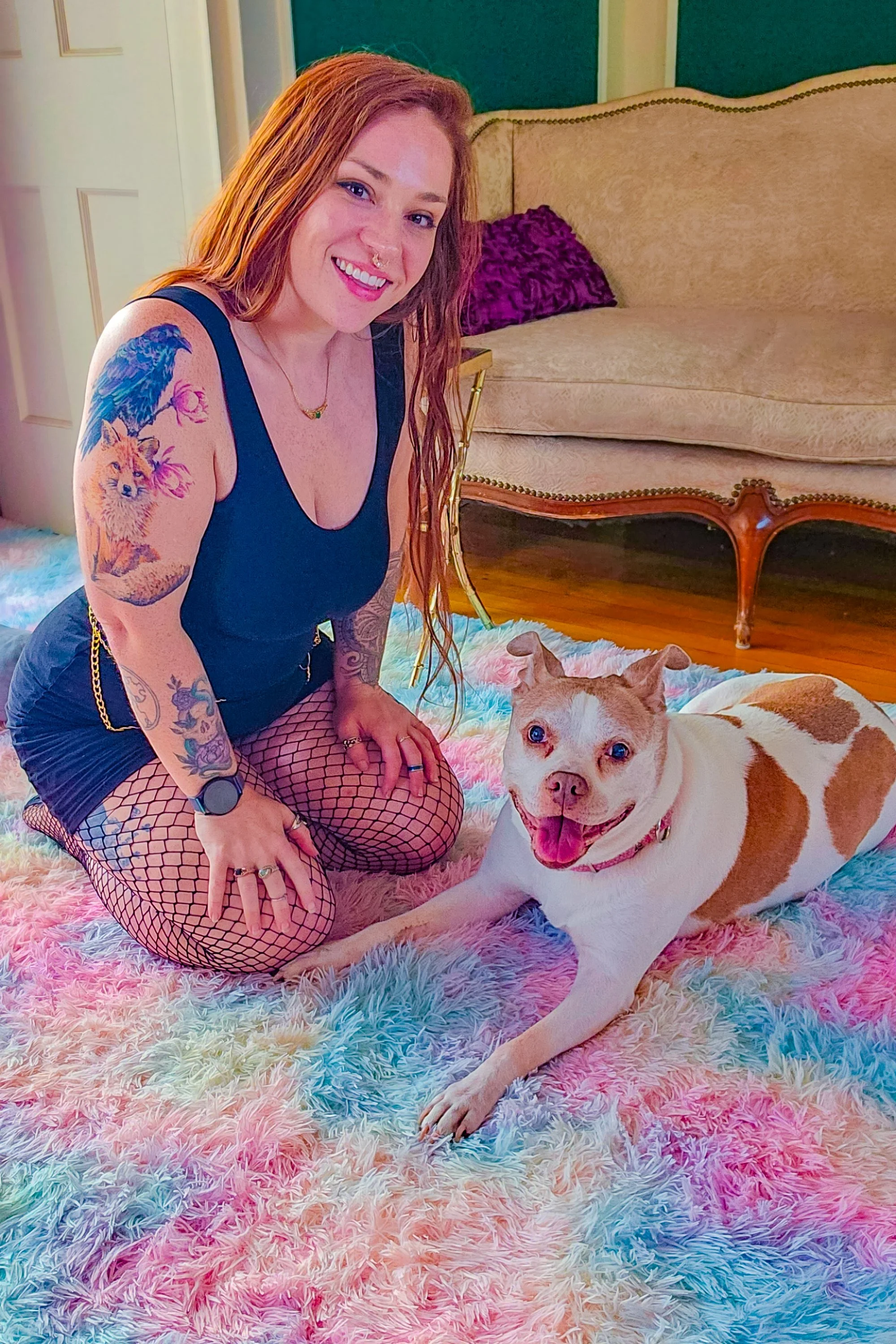 A woman with red hair, tattoos on her arm, wearing a black tank top and fishnet stockings, kneeling on a rainbow-colored fluffy rug, smiling next to a large white and brown dog lying on the rug. The woman is inside a room with a beige sofa and a purple pillow, with a teal and white wall in the background.