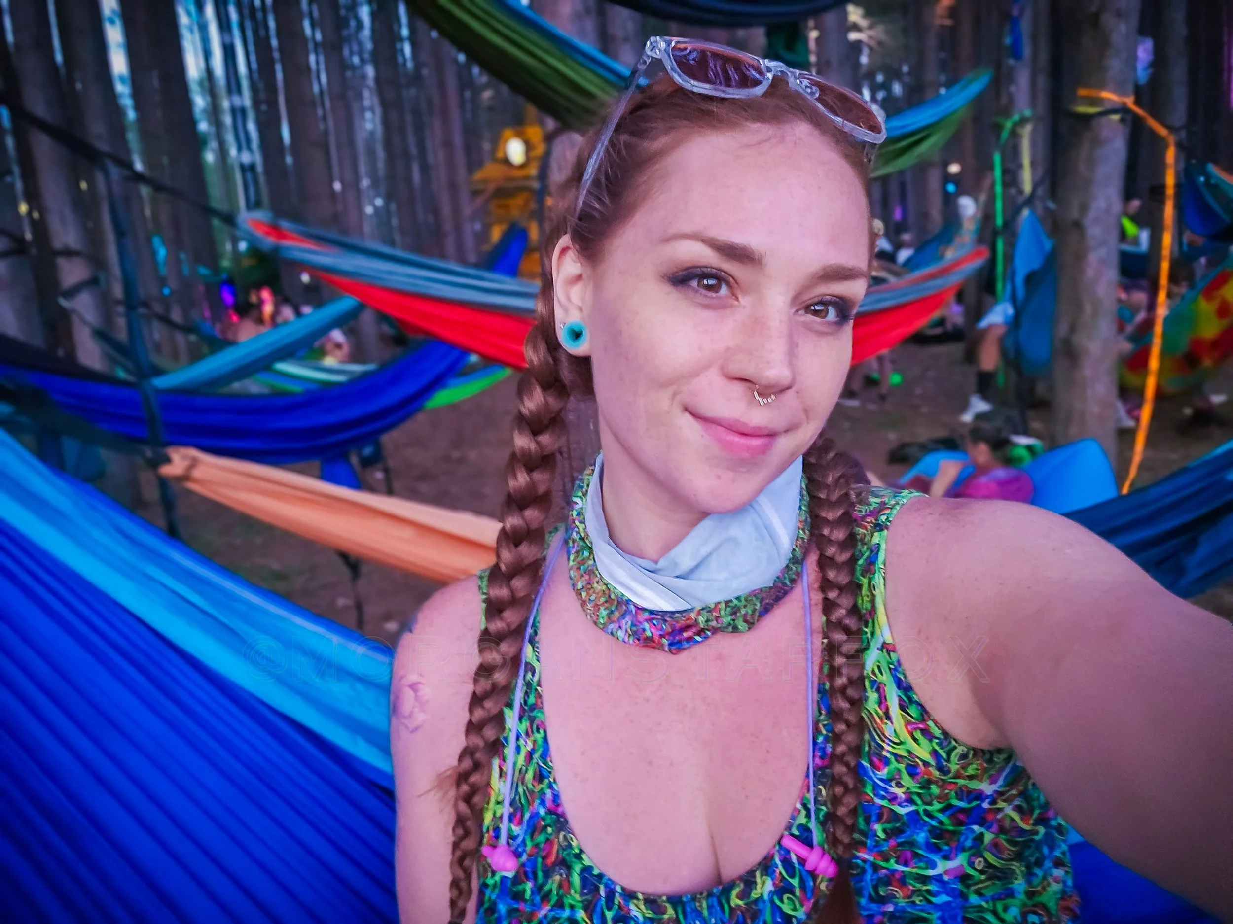 A girl with braided hair, sunglasses on her head, a nose ring, and turquoise earrings taking a selfie in a forest with colorful hammocks and people in the background.