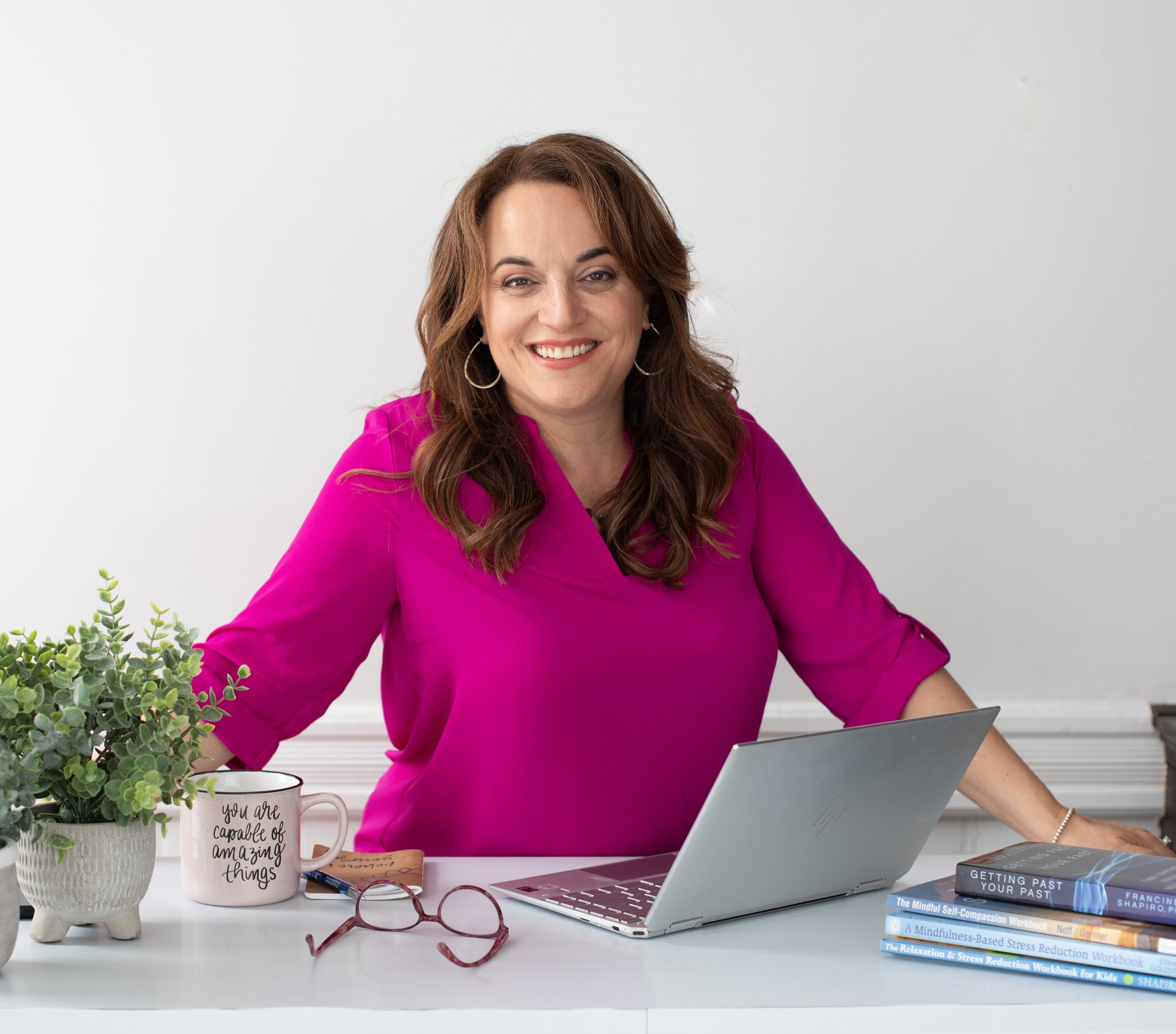Female therapist in Rhode Island smiling and standing in professional headshot with silver laptop, potted plant, mug with motivational text, pair of glasses, and counseling books.