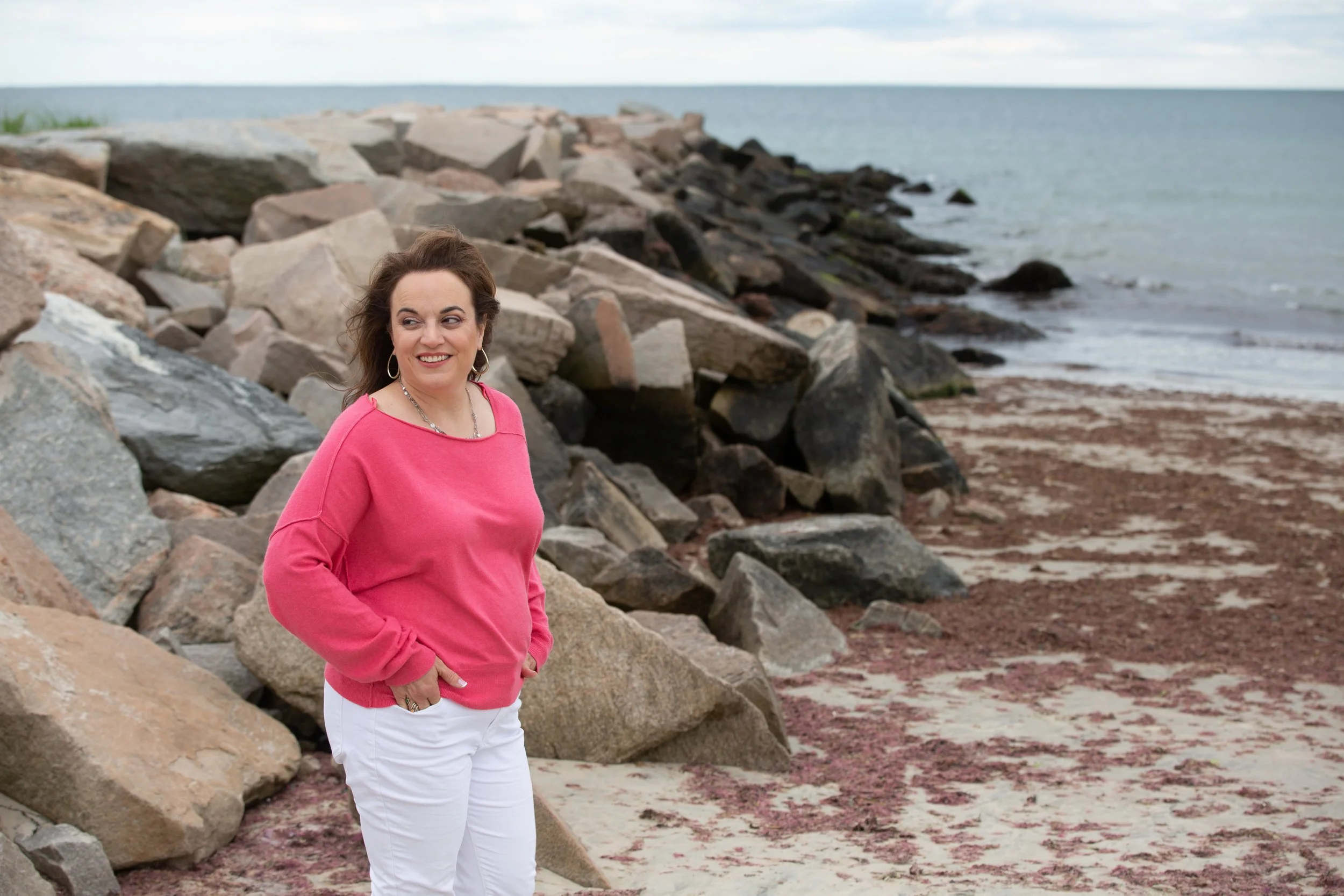 Woman counselor in pink sweater and white pants standing on rocky beach releasing anxiety in nature..