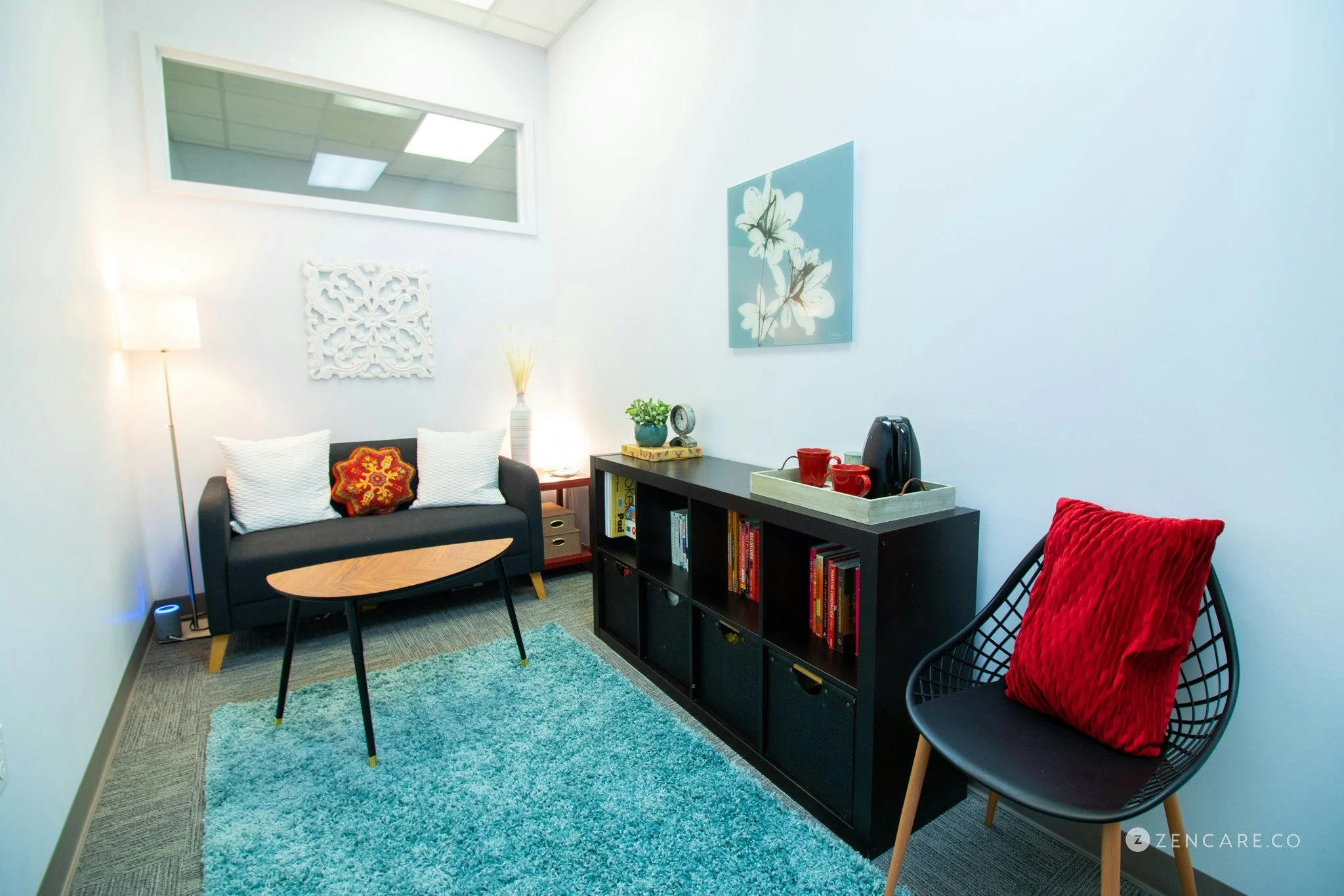 Cozy modern living room with a black sofa, white cushions, a colorful throw pillow, a small coffee table, a black chair with a red cushion, a blue rug, a black bookshelf with books and decor, a tray with cups and an electric kettle, framed artwork of flowers, a standing lamp, and a window high on the wall.