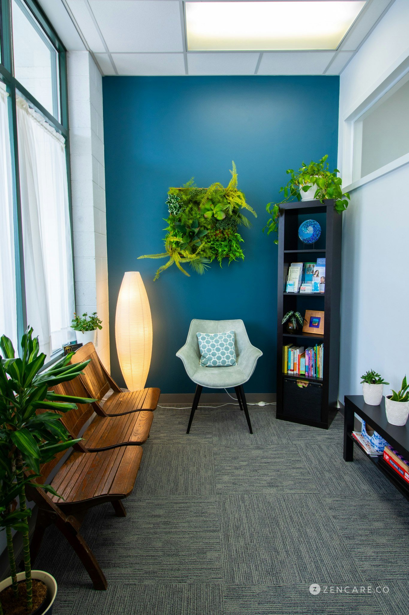 A waiting room with a blue accent wall, indoor plants, a wooden bench, a white armchair with a patterned pillow, a tall floor lamp, a black bookshelf with decorative items and books, and large windows with white curtains.
