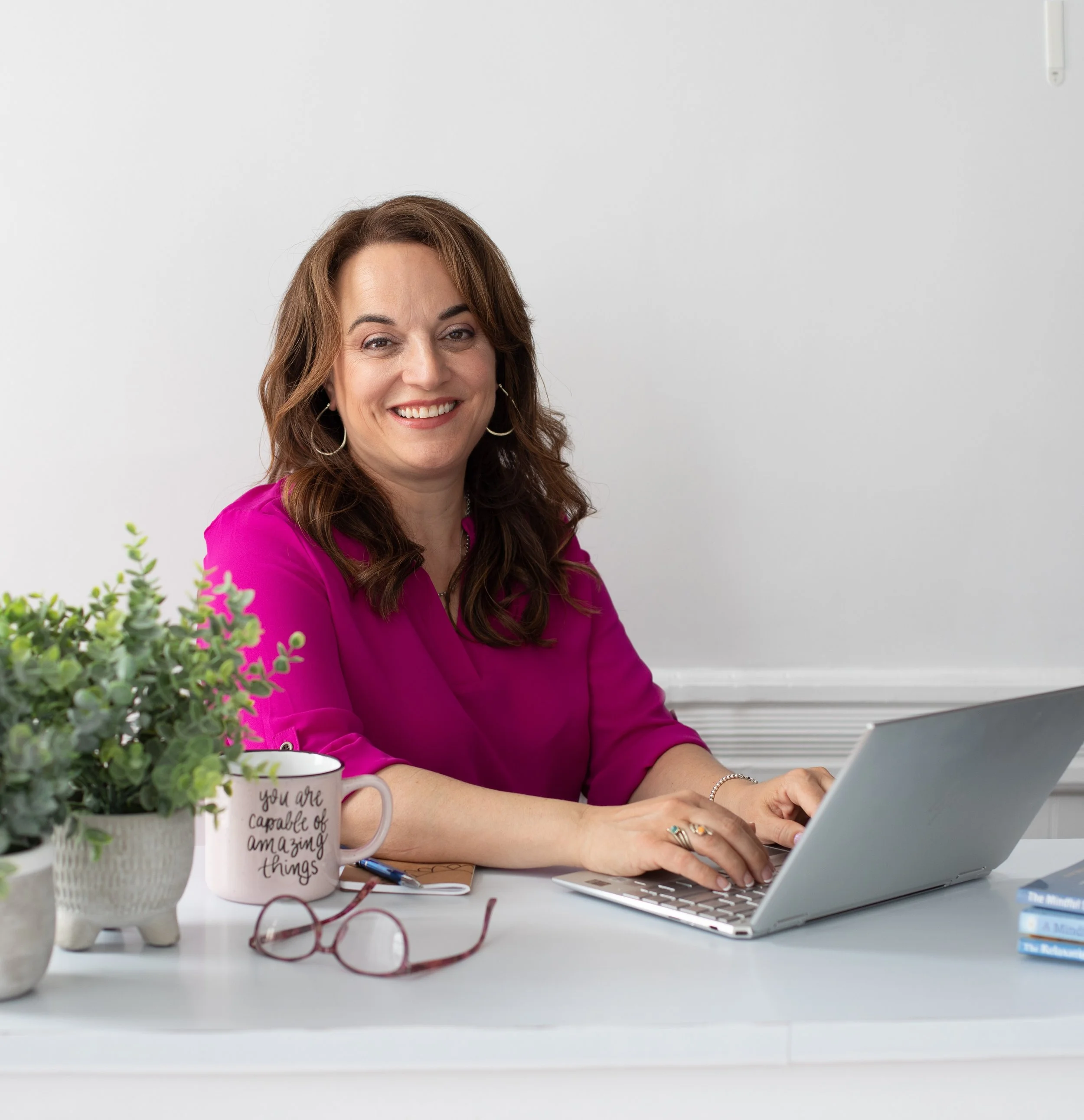 Woman mental wellness coach wearing pink, smiling while working on laptop at desk. On desk are potted plant, pink mug with motivational mantra, pair of glasses, and mindful self-compassion books.