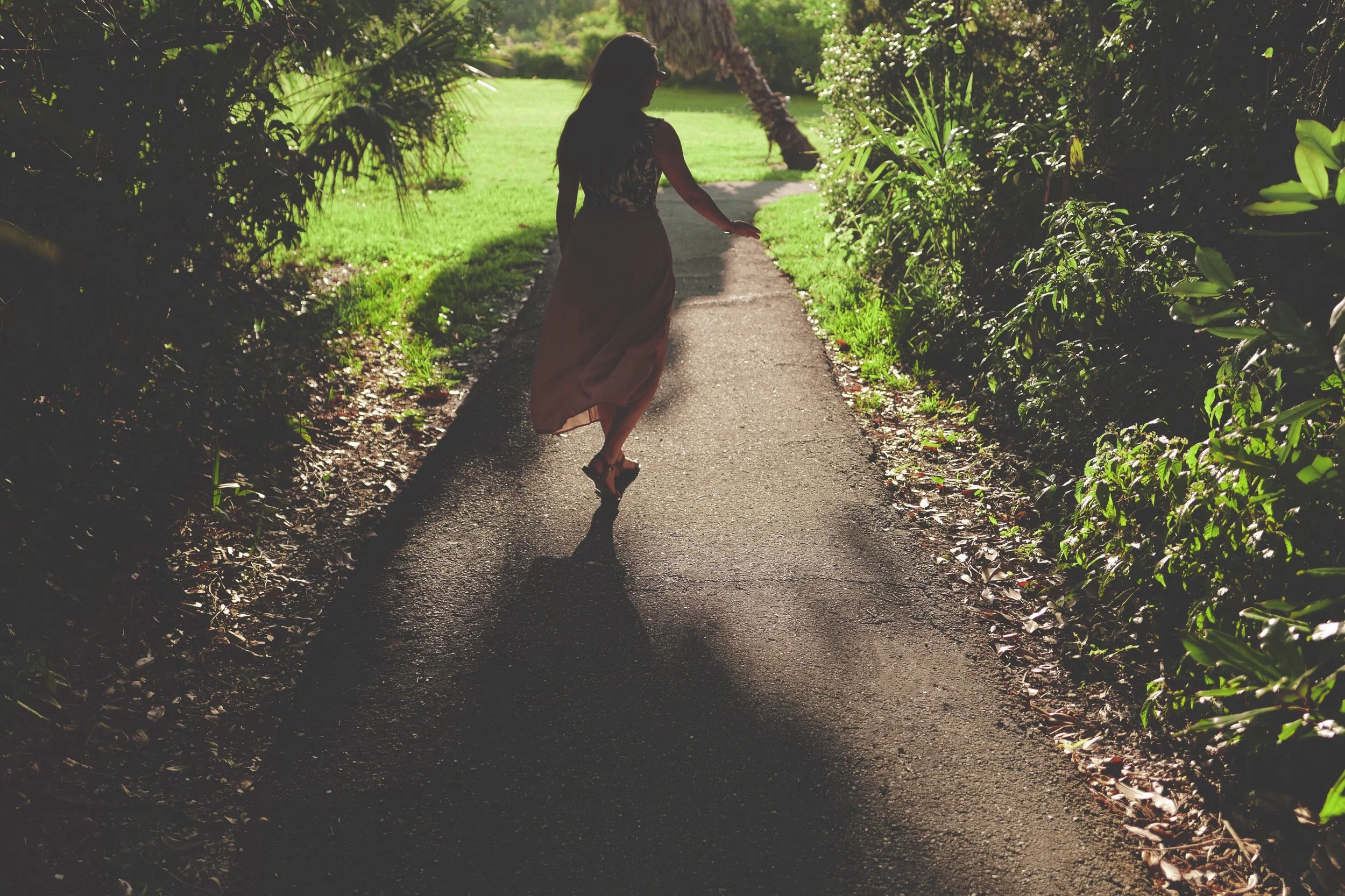 Woman in long dress walking in mindfulness on sunny, shaded pathway surrounded by green plants and trees.