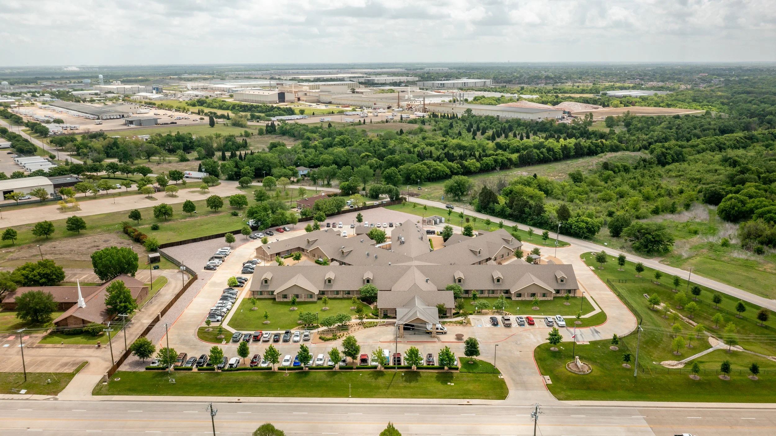 Aerial view of a large building complex with surrounding parking lots, green trees, and open fields, with an industrial area in the background.