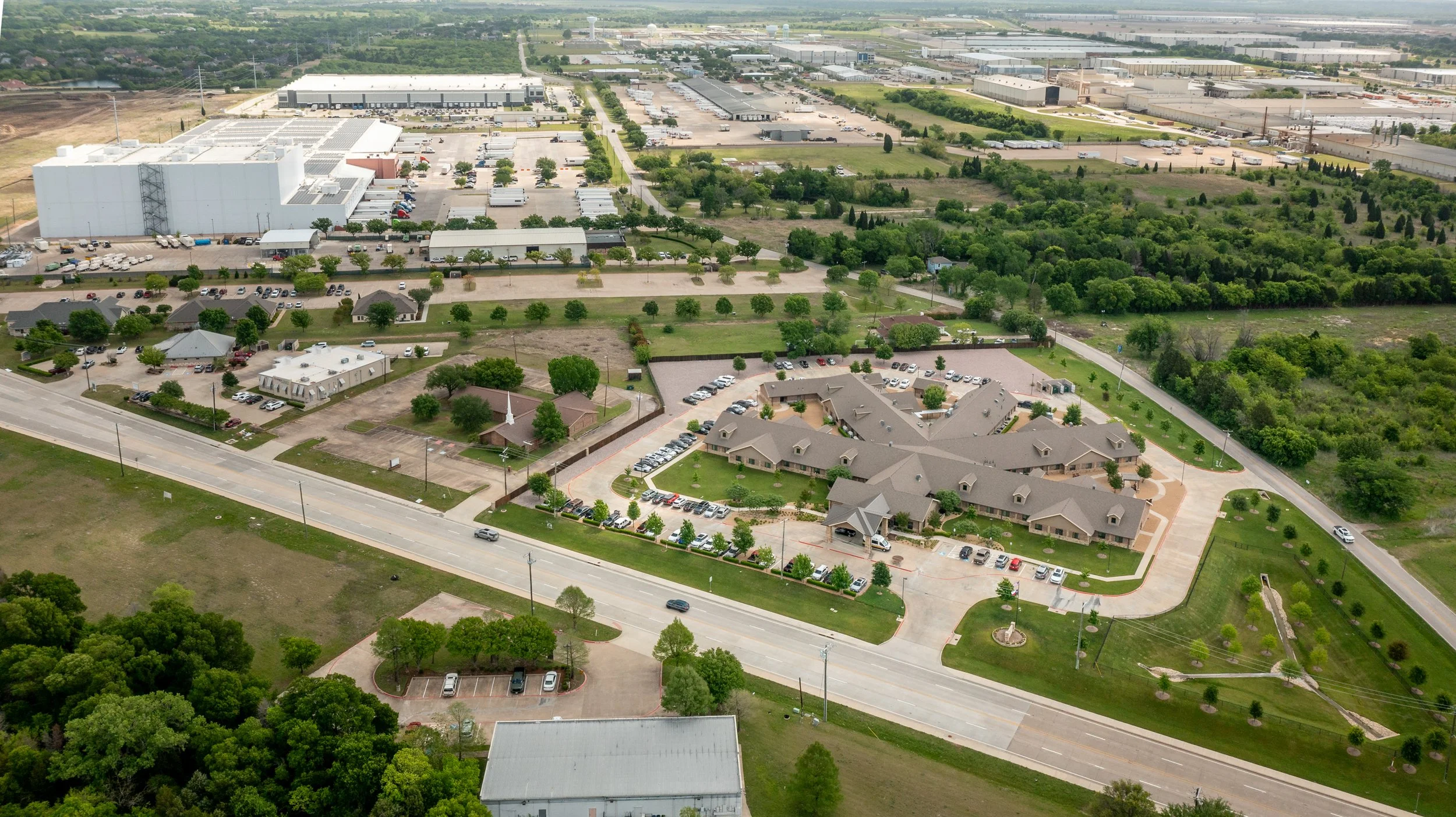 An aerial view of a mixed-use area with residential buildings, commercial or industrial warehouses, parking lots, and green spaces with trees and grass fields.