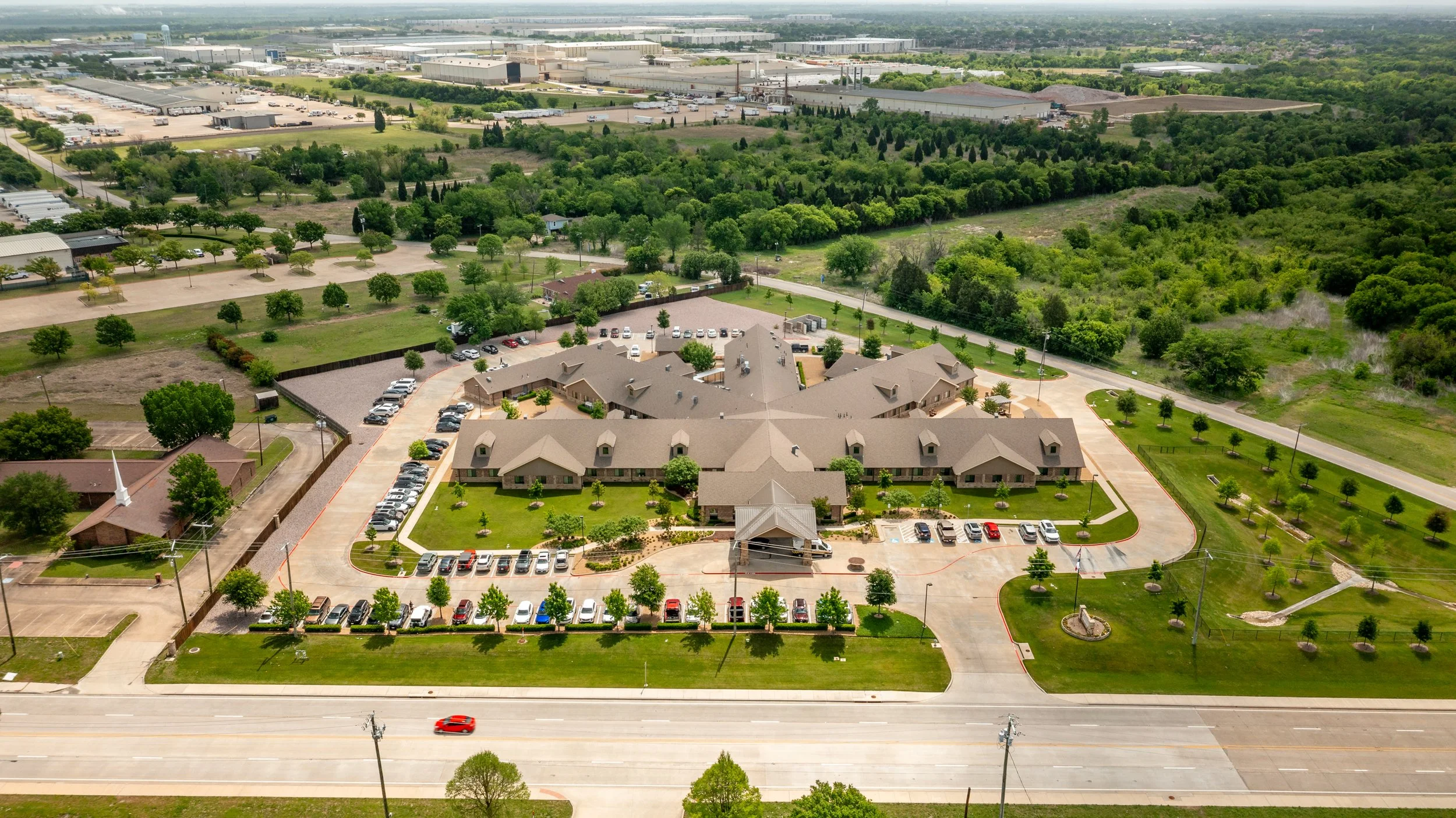 An aerial view of a building complex with a parking lot, surrounded by greenery and trees, with an industrial area in the background.