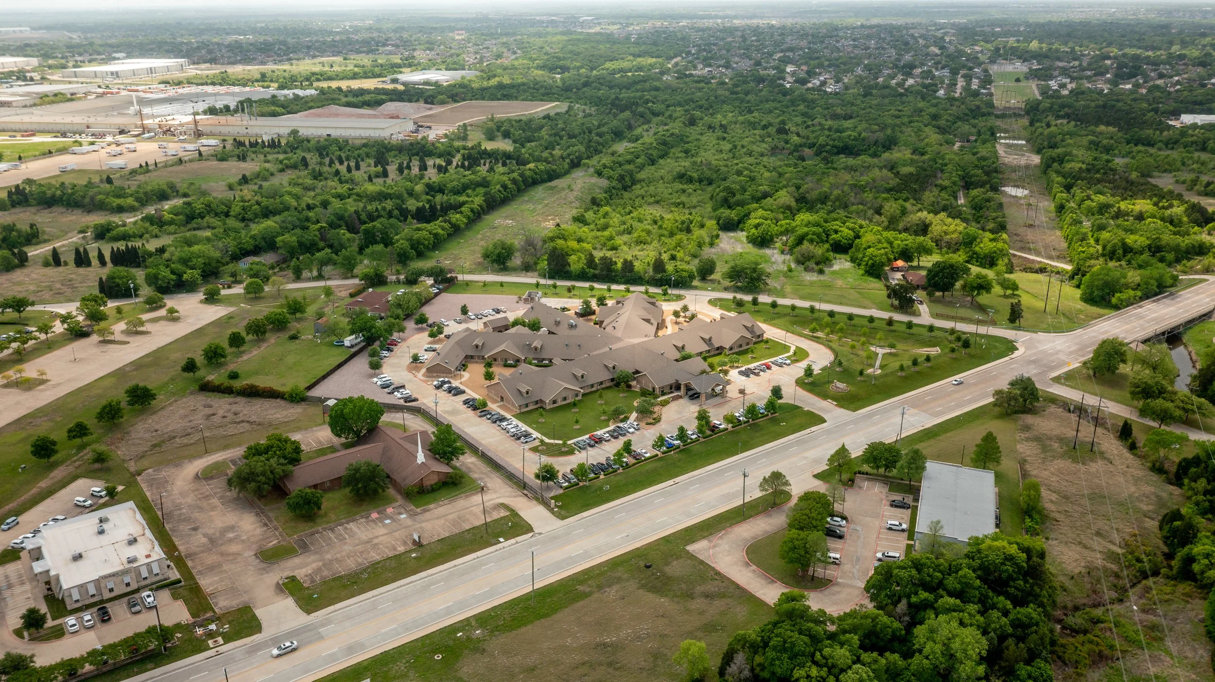 An aerial view of a suburban area with a mix of residential buildings, parking lots, green spaces, and wooded areas, with a highway running through the bottom of the image.
