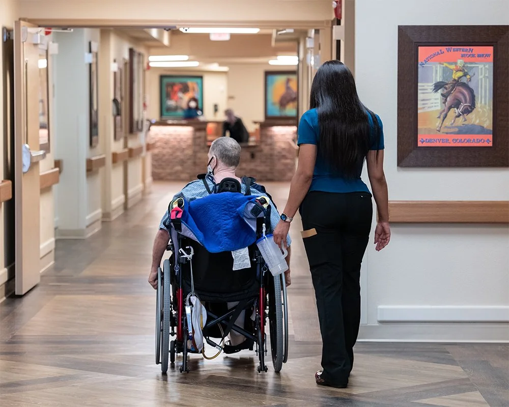 A healthcare worker assists a person in a wheelchair down a hallway in a medical facility.