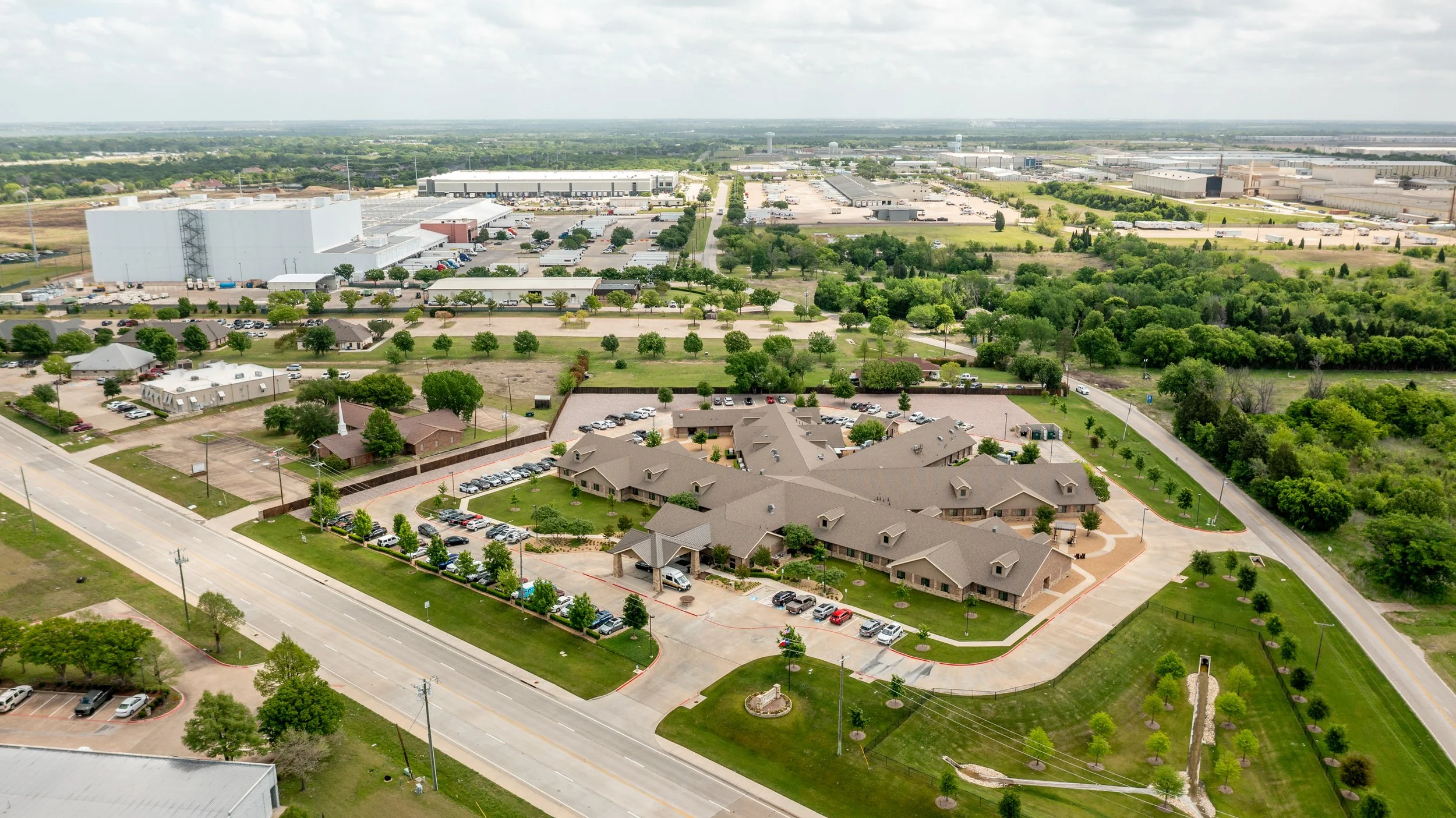 An aerial view of a suburban residential area with rows of houses, parking lots, trees, and green spaces, transitioning into an industrial area with warehouses, large buildings, and parking lots in the background.