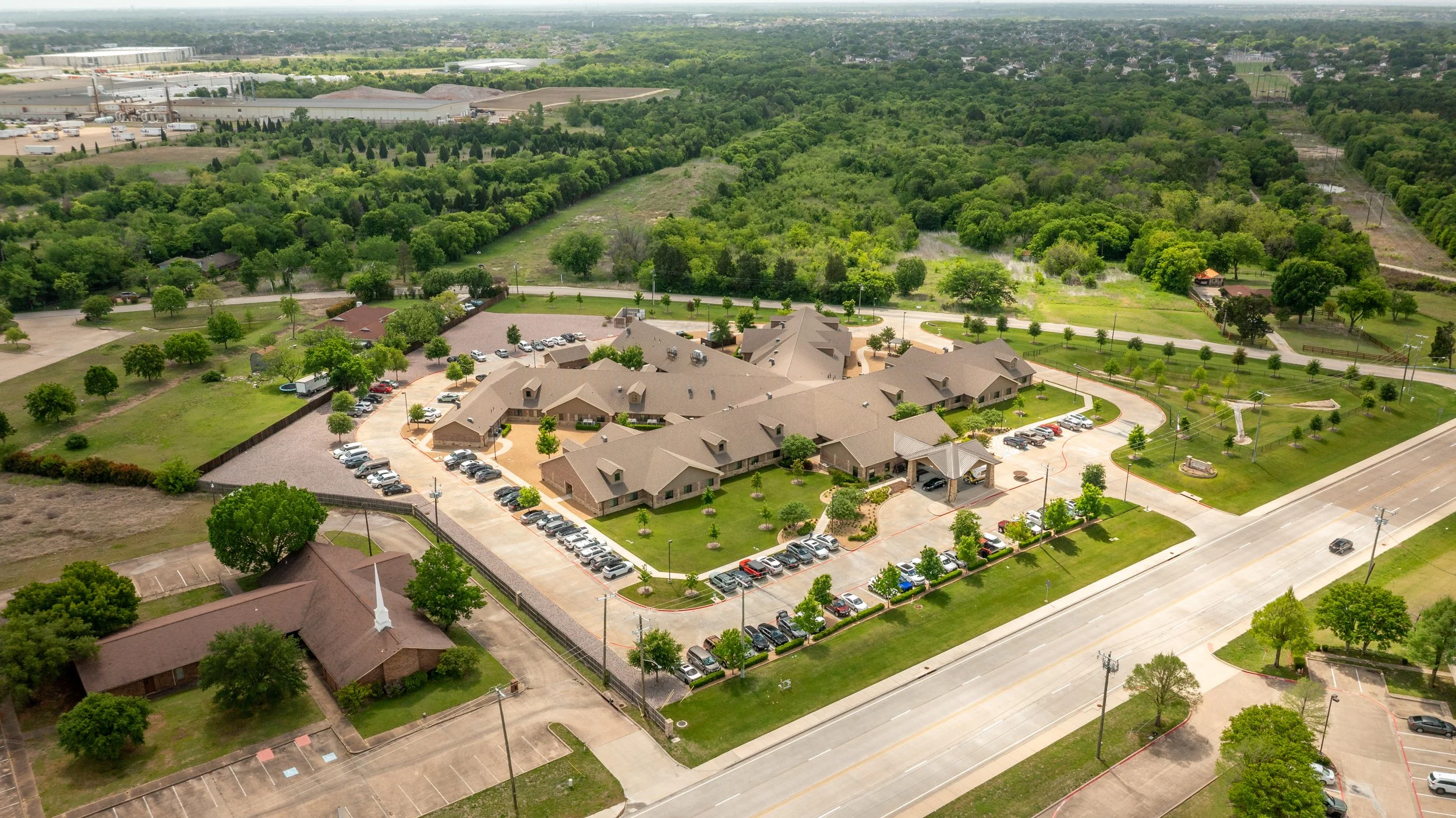 Aerial view of a residential complex with beige buildings, parking lots with cars, surrounded by green trees and open fields.