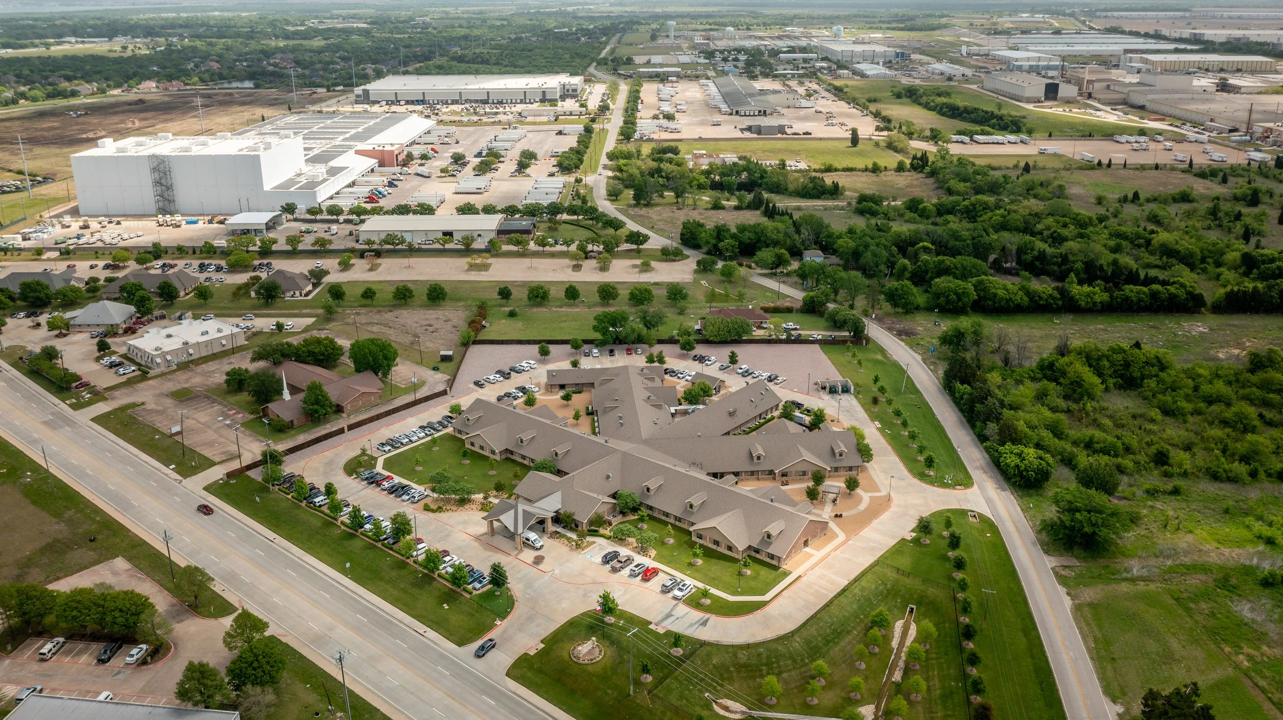 Aerial view of a mixed residential and industrial area showing houses, parking lots, large warehouses, and green spaces with trees, roads, and open fields.