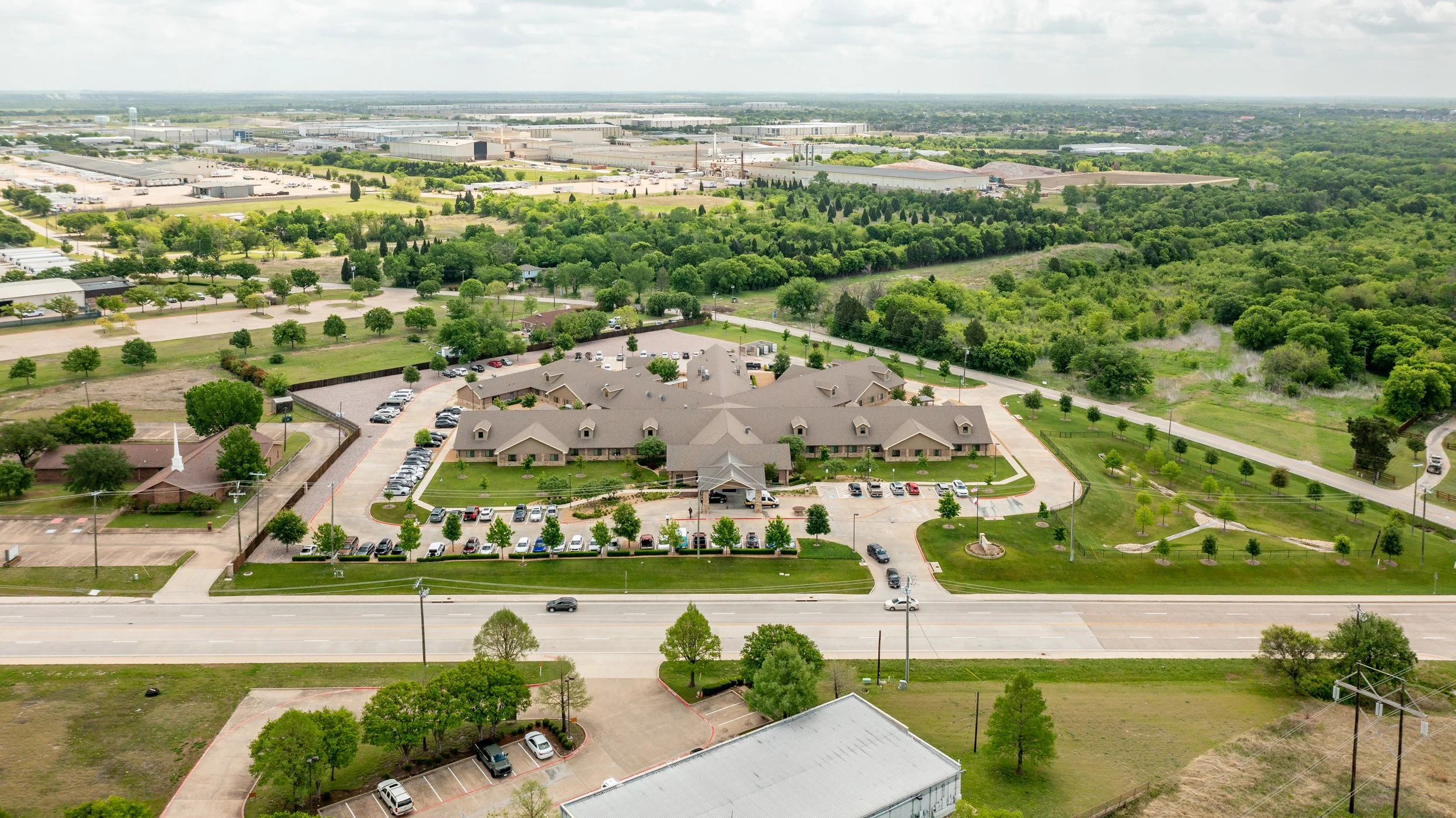 Aerial view of a mixed-use area with residential homes, a commercial building, parking lots, and a lush green forest in the background.